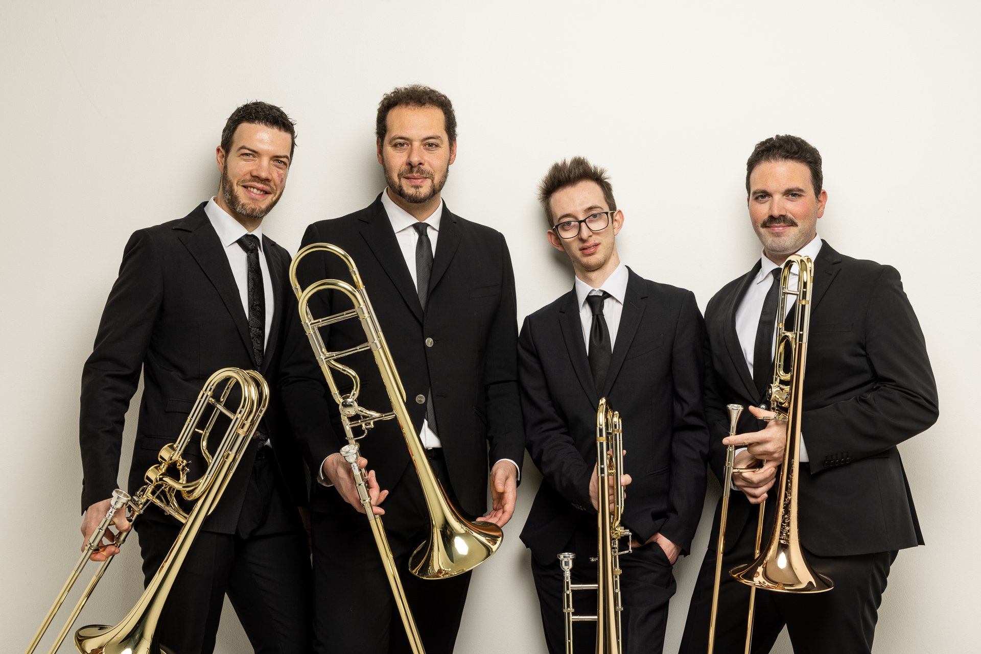 Un gruppo di cinque uomini in abiti eleganti con strumenti musicali a fiato, posizionati contro una parete bianca.