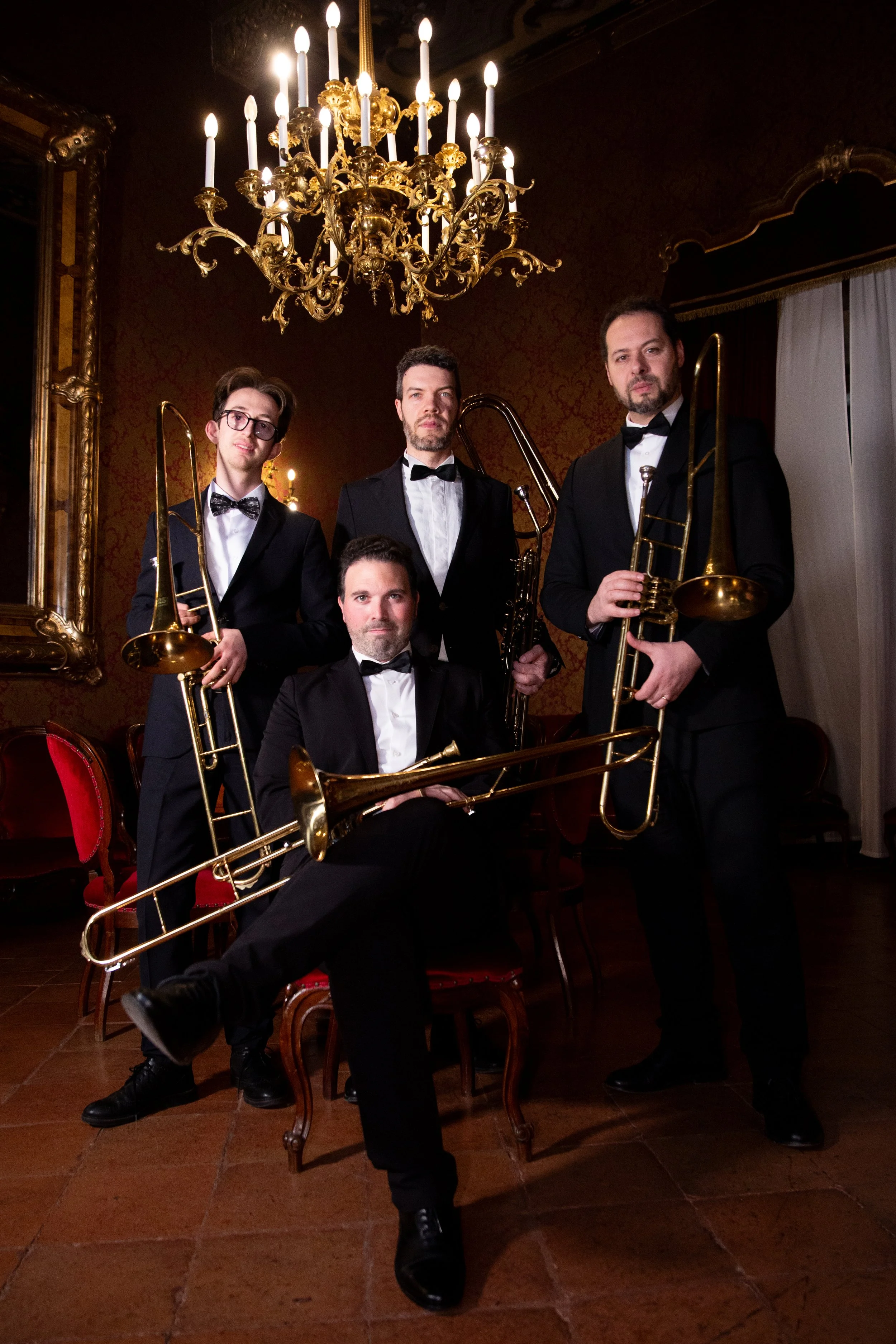 Un gruppo di cinque uomini vestiti con tuxedo, alcuni con strumenti musicali a fianco, in un ambiente elegante con lampadario dorato e pareti decorate.