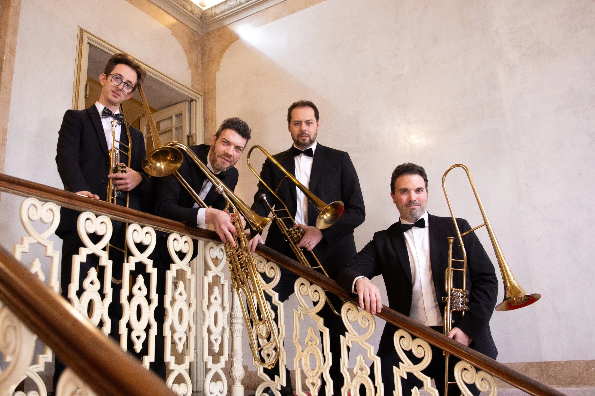 Cinque uomini in elegante abbigliamento formale con giacche nere e papillon, alcuni con strumenti musicali come tromboni e un trombone, posano sulla ringhiera di una scala interna in un edificio con pareti beige e dettagli decorativi.