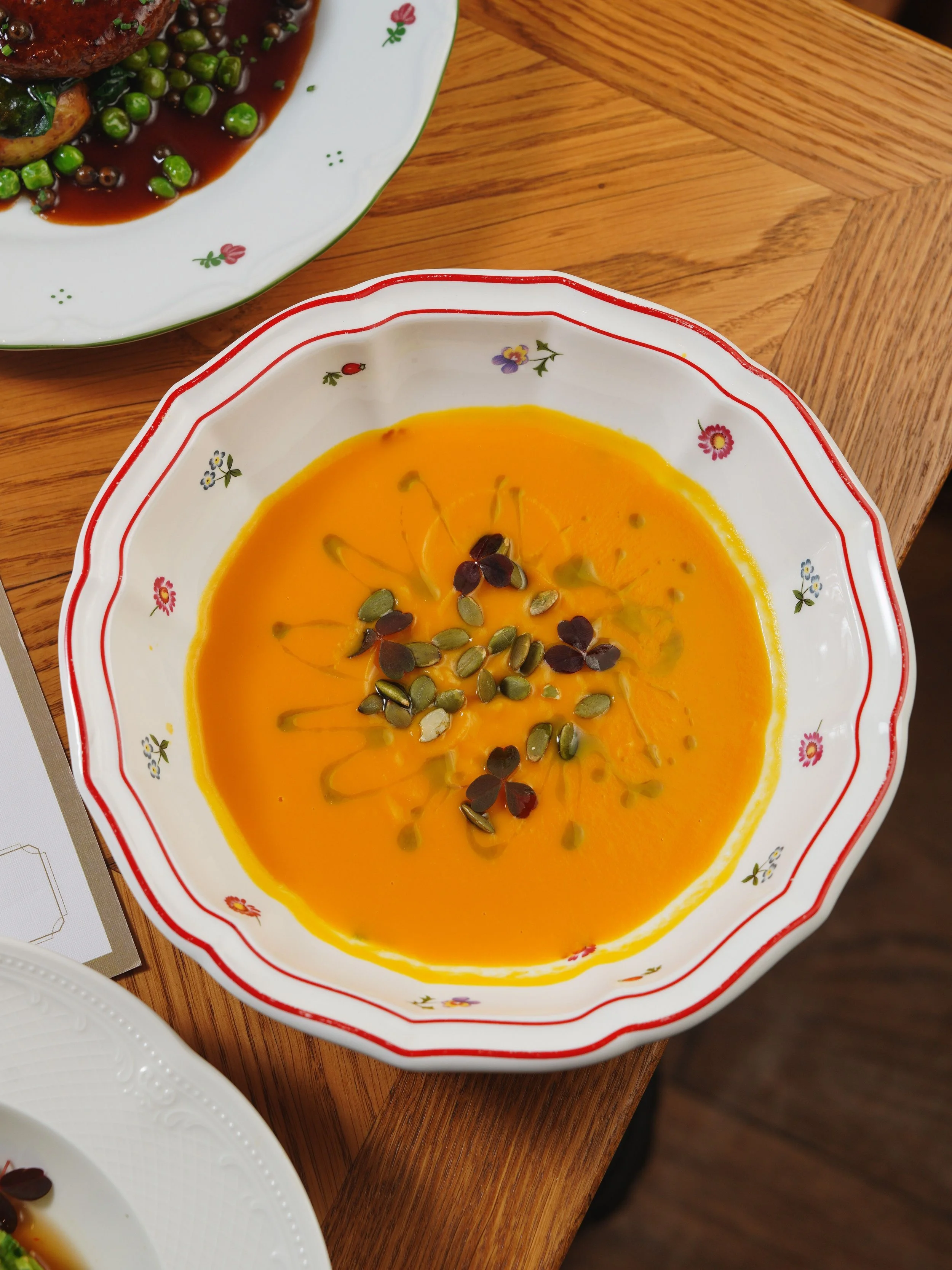 Orange squash soup garnished with pumpkin seeds and small red-purple microgreens in a patterned bowl