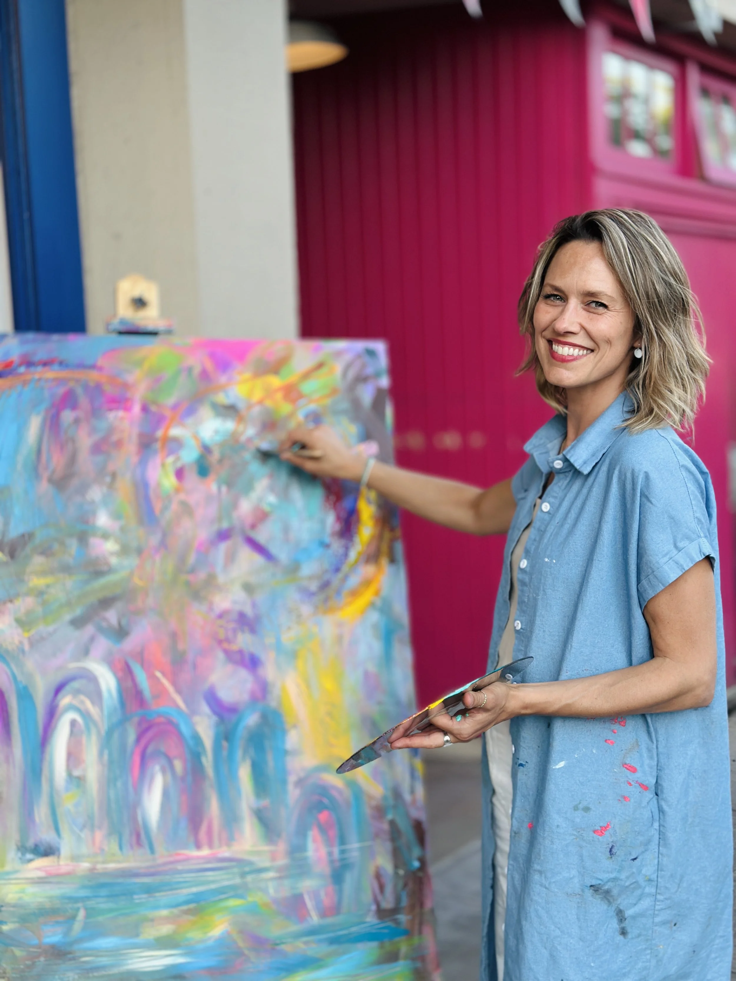 A vertical photo of an artist in a colorful studio setting. She is standing behind a brightly colored abstract painting that features arched, rainbow-like strokes in blues, pinks, and yellows