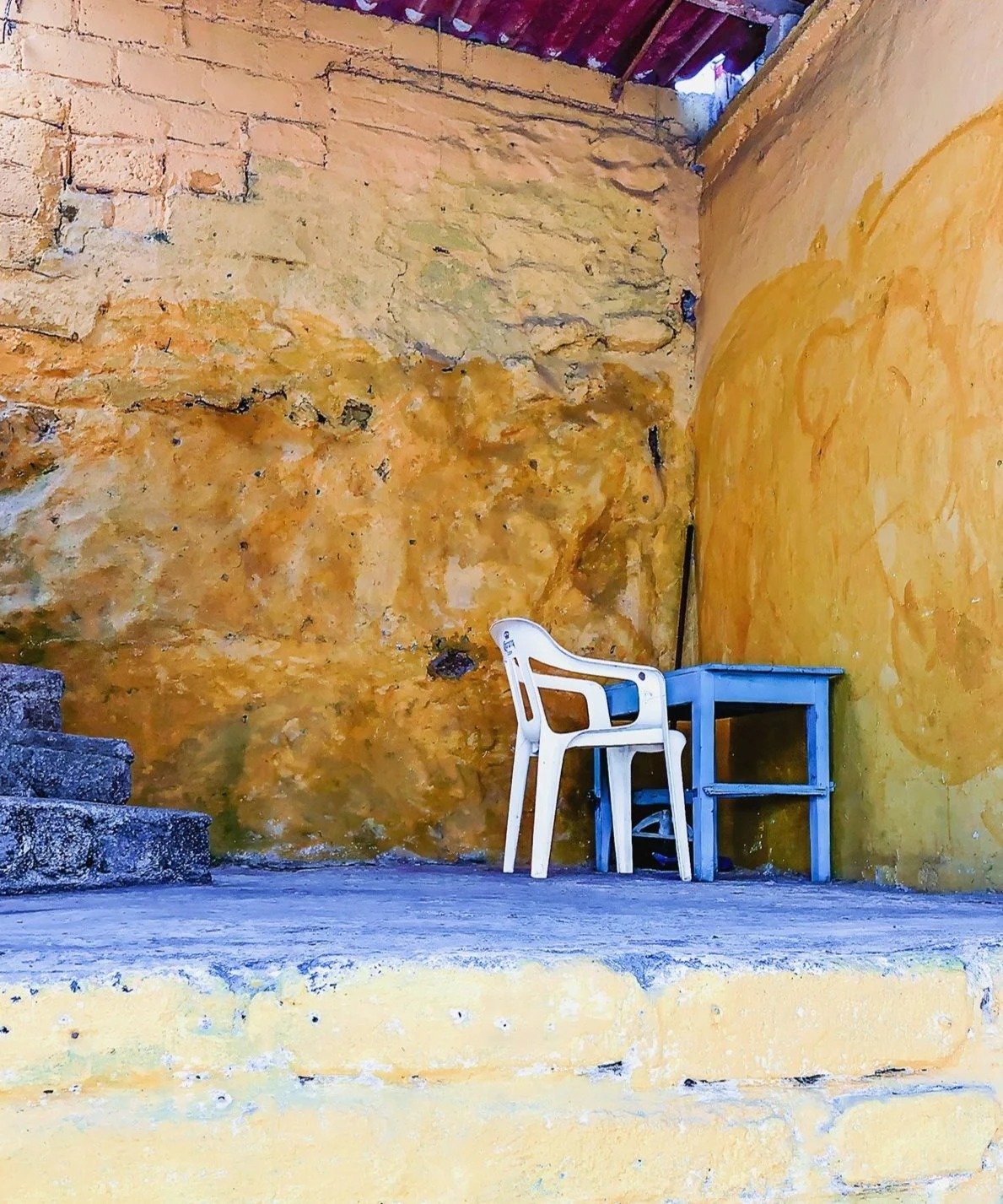 An outdoor area with a textured yellow and orange wall, a white plastic chair, a small blue table, and stone steps.