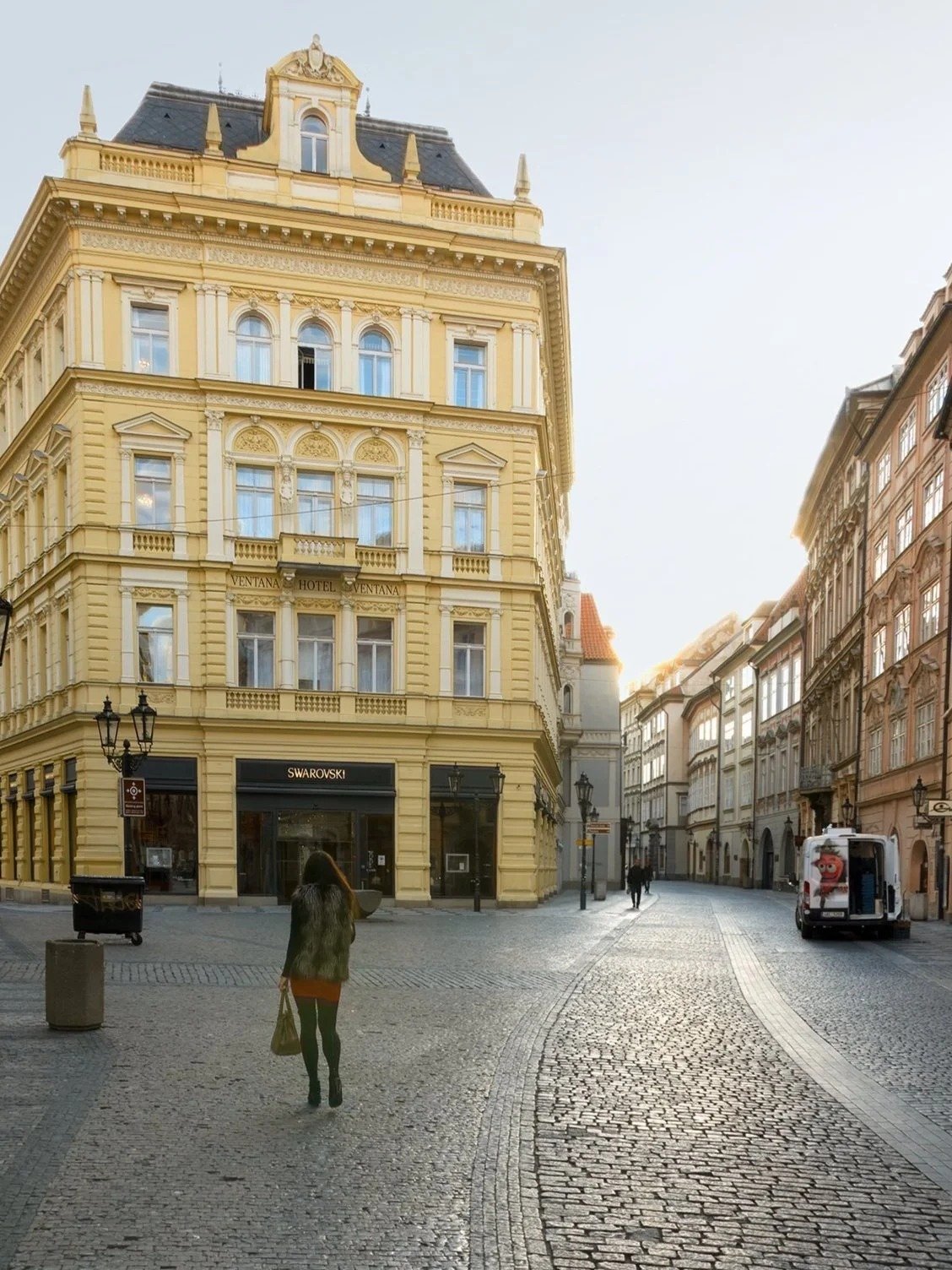 A woman walking on a cobblestone street in front of a yellow historic building with a Swarovski store on the ground floor, during sunset in a European city.