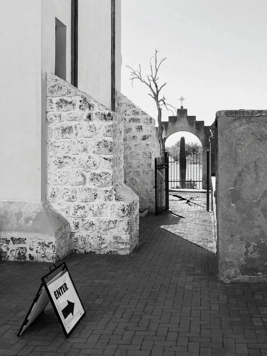 Black and white photo of a stone archway with a gate, a leafless tree, and a sign on the ground with an arrow pointing right that says 'ENTER'.