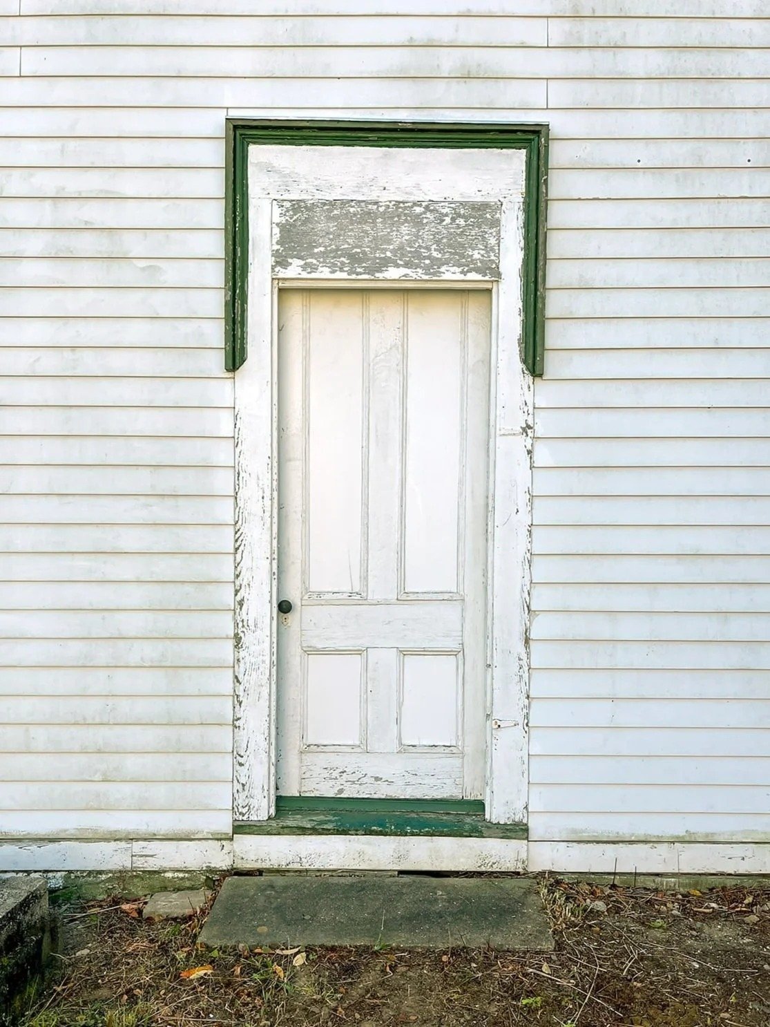 A weathered white wooden door with peeling paint, set in an old white siding house, with a green trim frame and a small concrete step at the entrance.