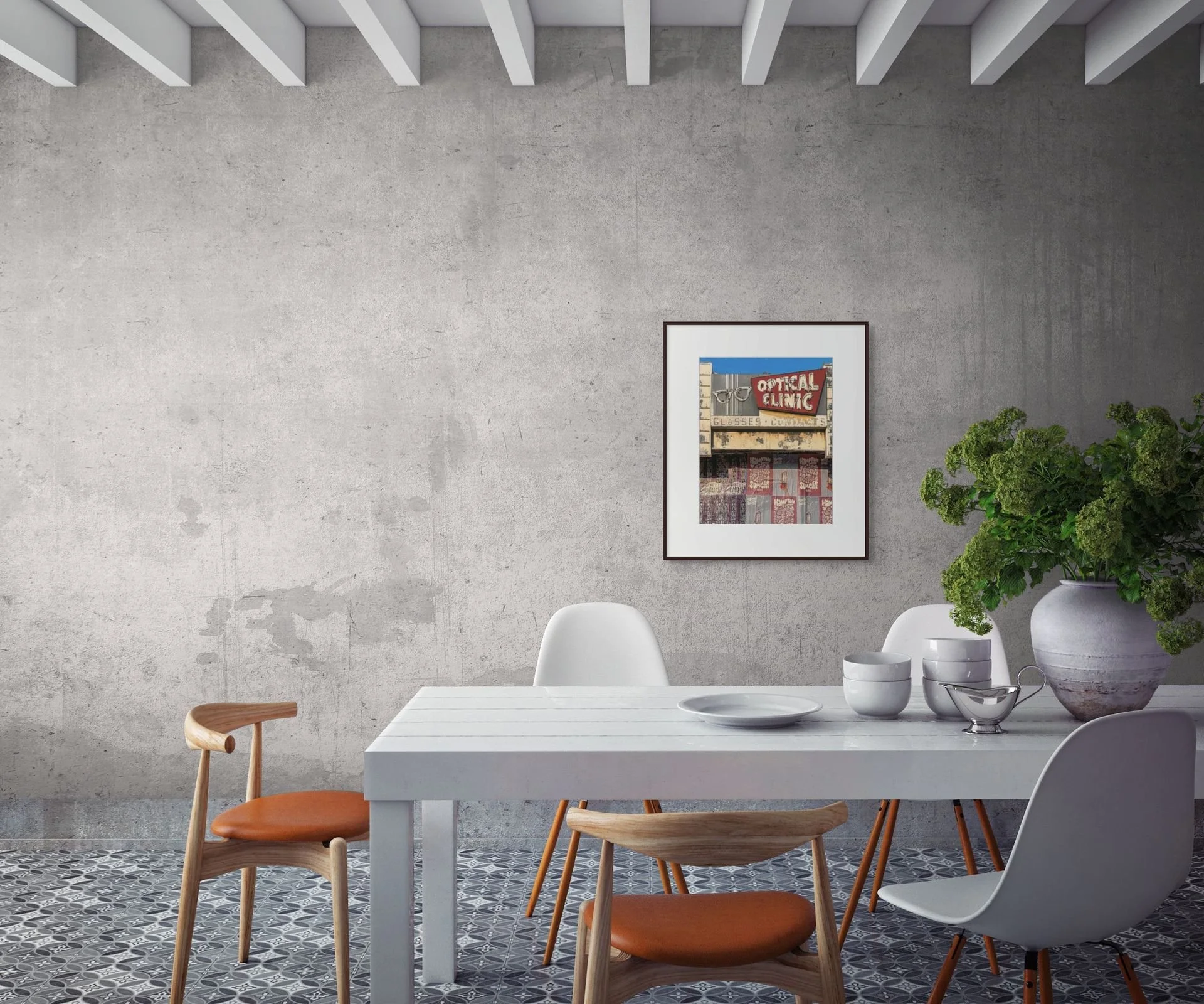 Modern dining room with white table, wooden chairs, gray concrete wall, framed artwork, large green plant in a white pot, and patterned tile floor.