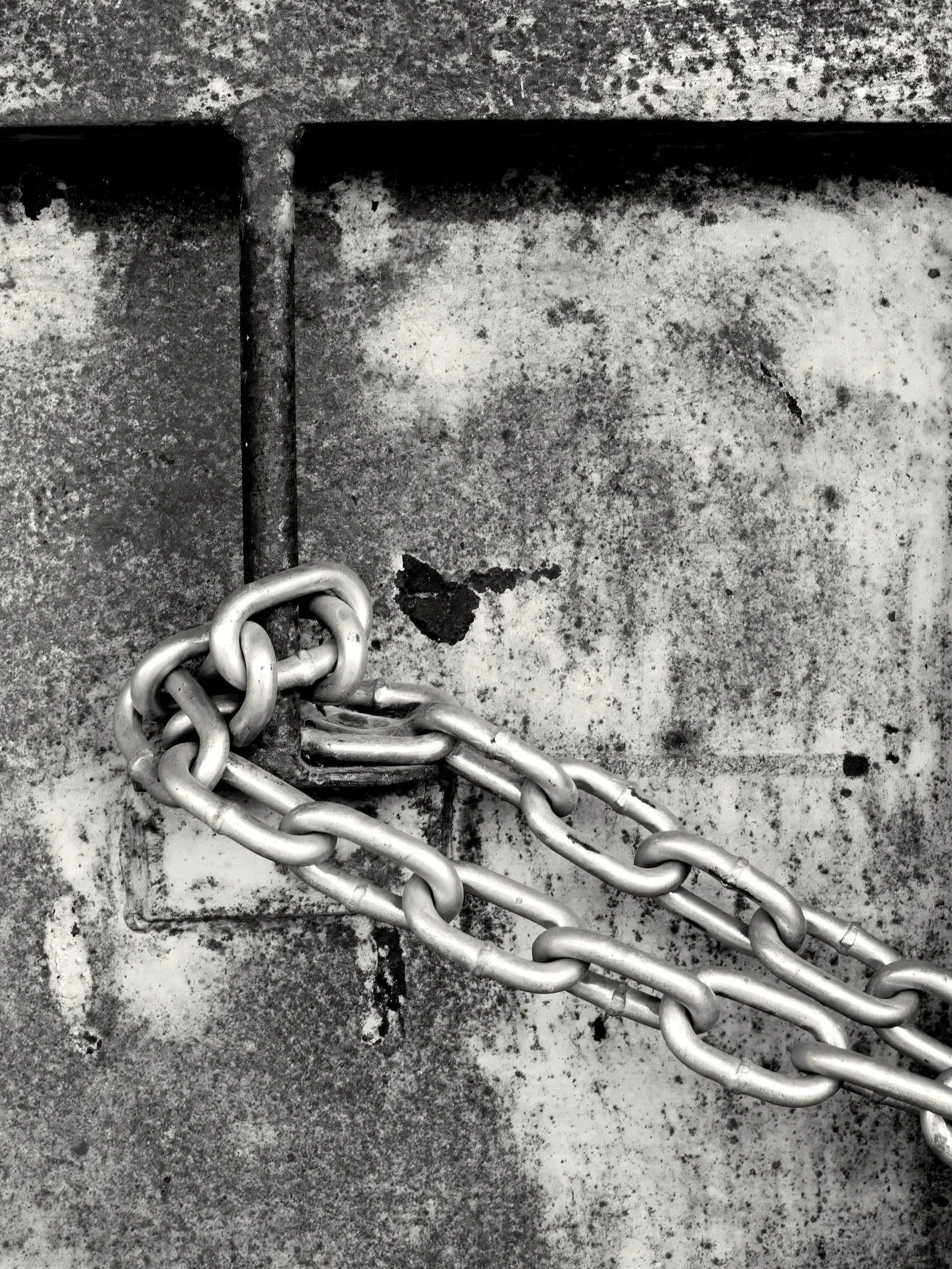 A close-up black and white photograph of a metal chain securing a locked metal fence, with the chain wrapping around a vertical bar and the fence showing signs of rust and wear.