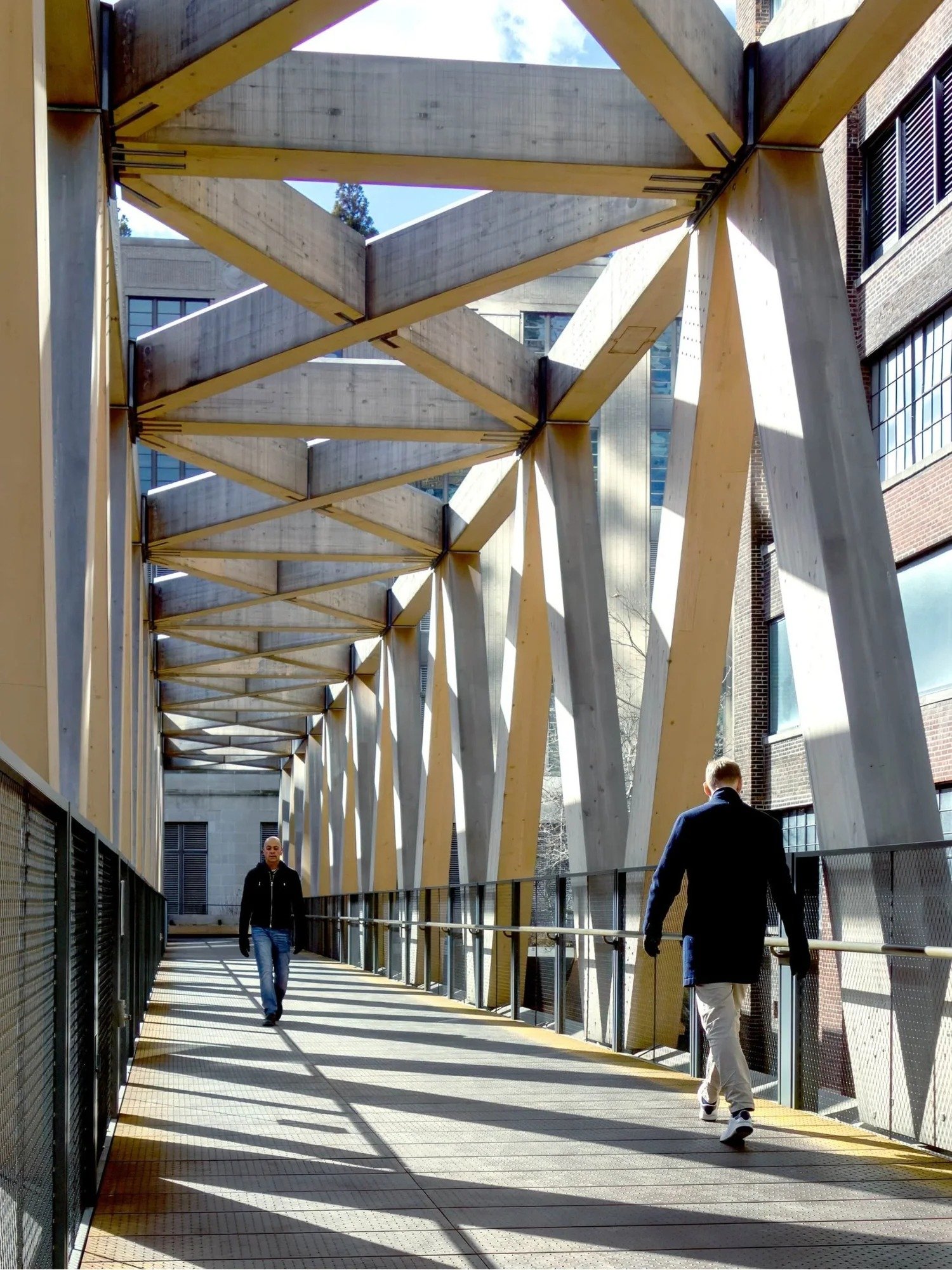 People walking on a modern outdoor walkway with wooden beams overhead and a metal railing, surrounded by tall buildings.