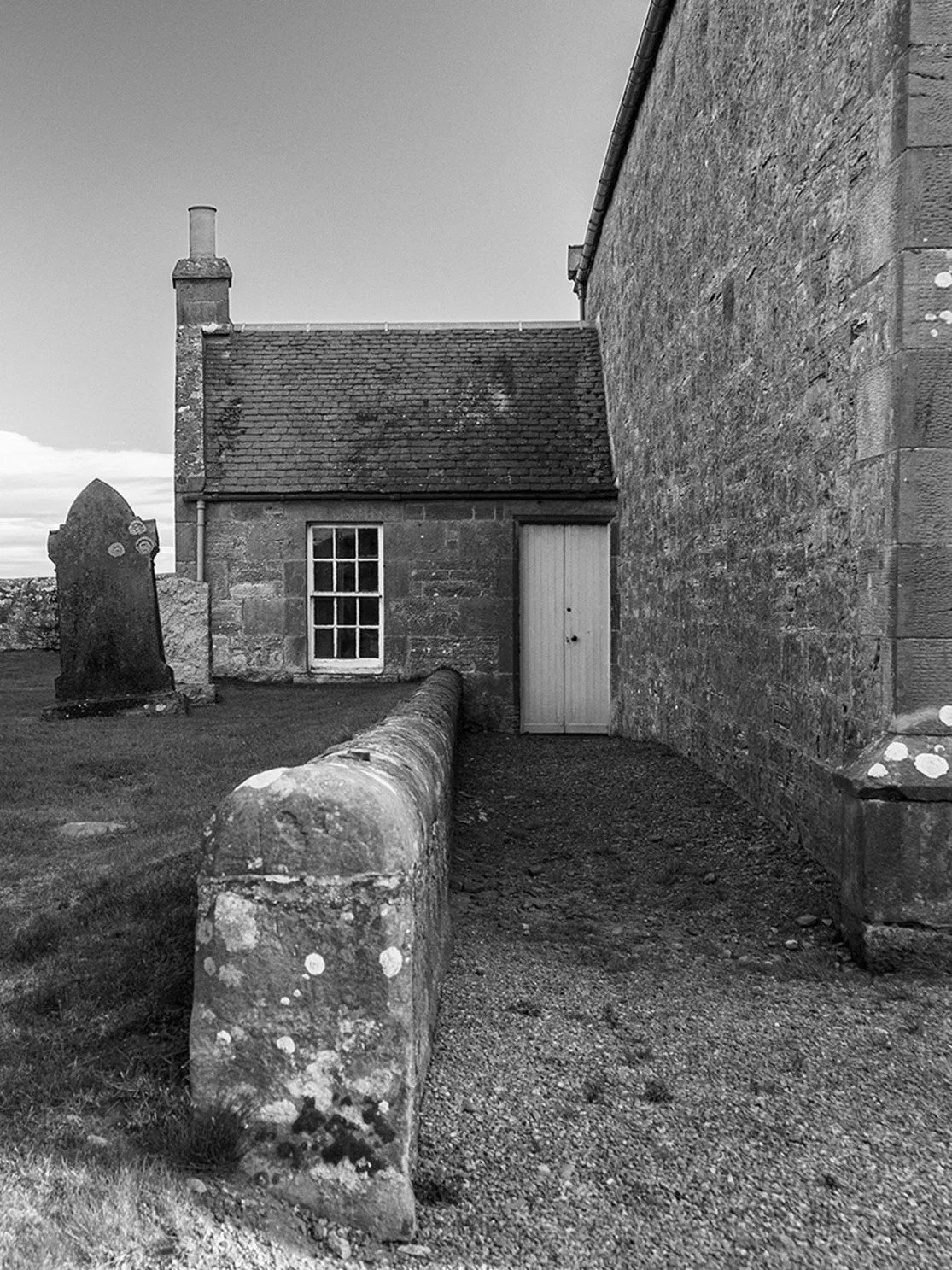Black and white photo of an old stone building with a small window, a door, and a chimney. There is a stone wall and a gravestone nearby.