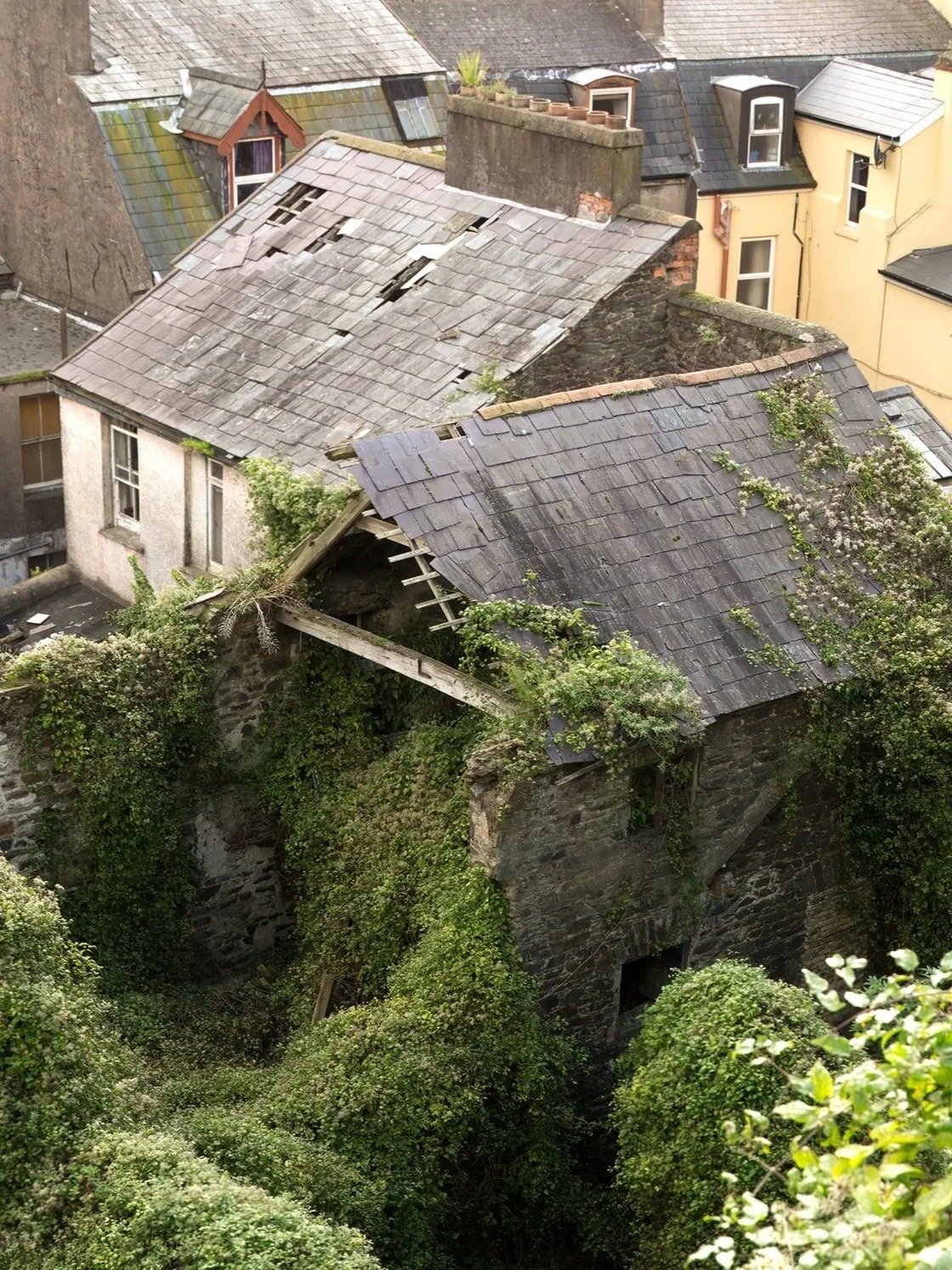 An old, abandoned building with a damaged roof, covered in greenery and vines, surrounded by other houses.