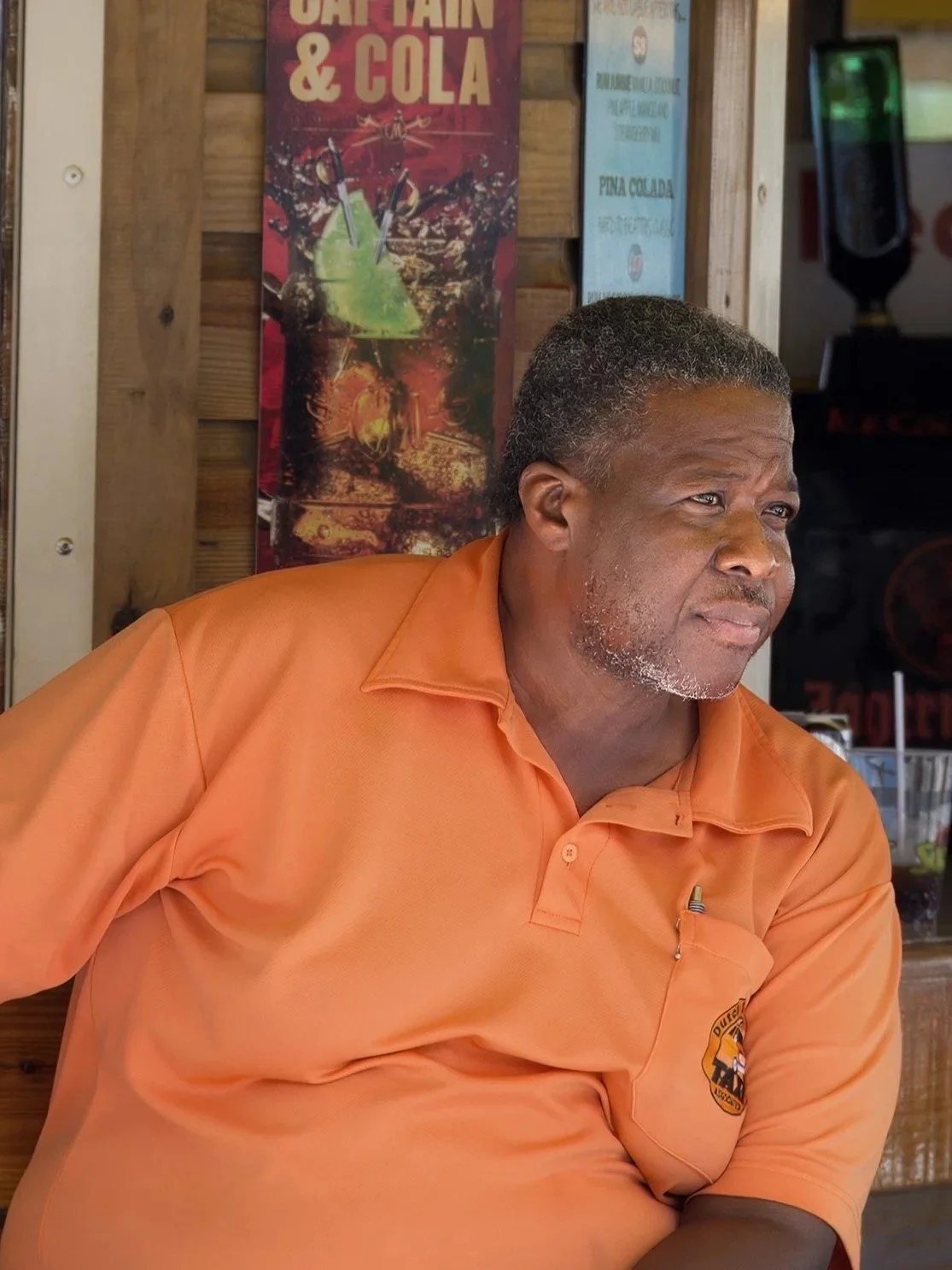 A man with short gray hair and a beard wearing an orange polo shirt with a logo on the left chest, sitting inside a bar or restaurant setting.