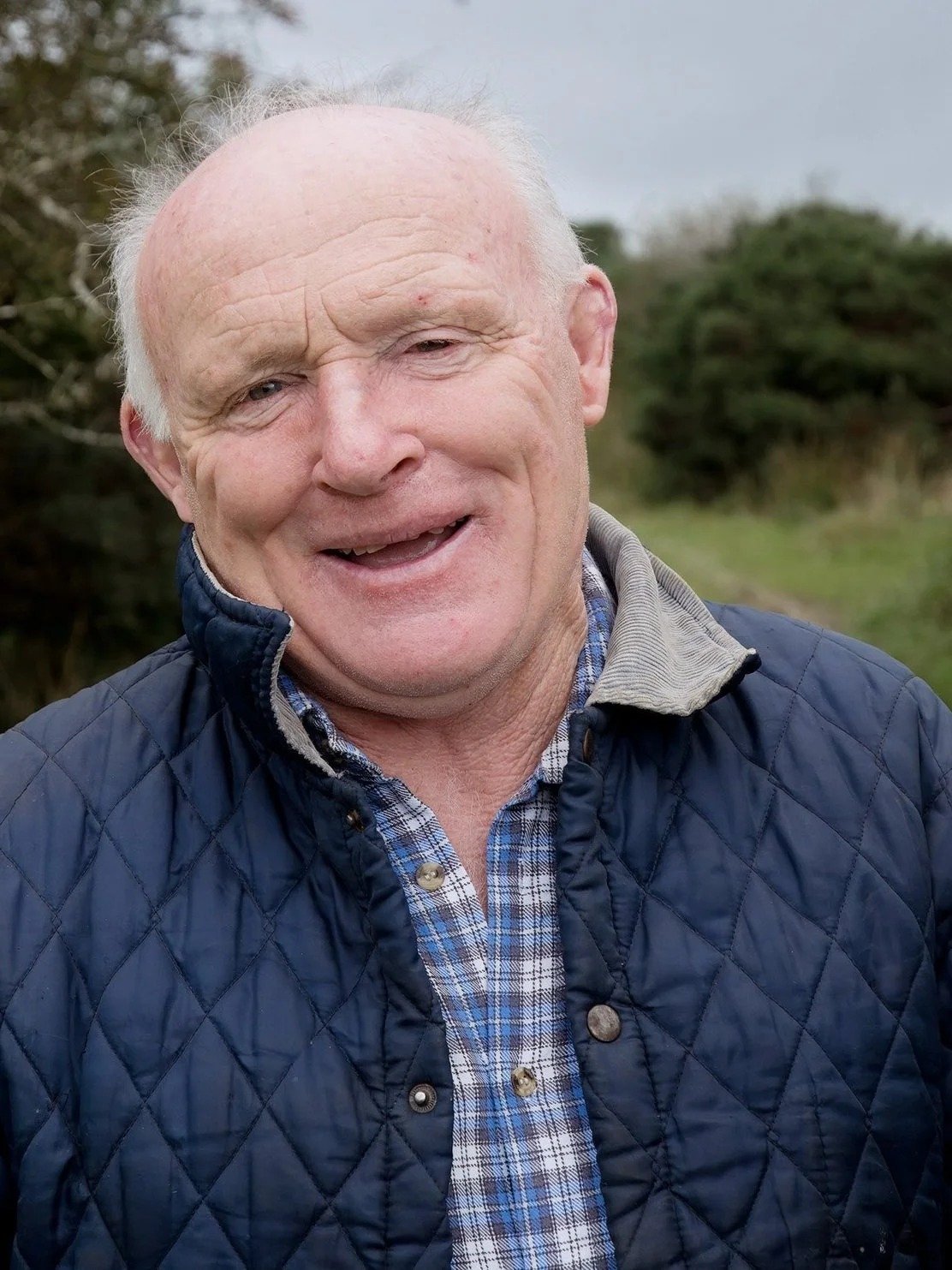 Close-up of smiling elderly man outdoors, wearing a navy quilted jacket over a plaid shirt, with greenery in the background.