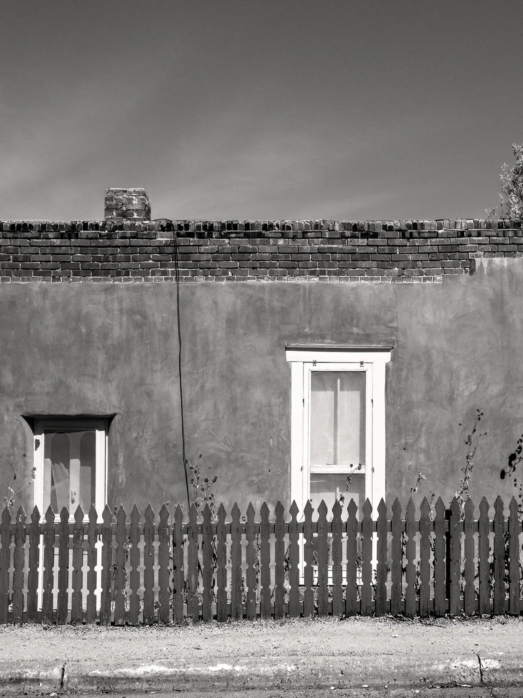 Black and white photo of a house with a brick chimney, two windows with white trim, a wooden fence, and a clear sky.