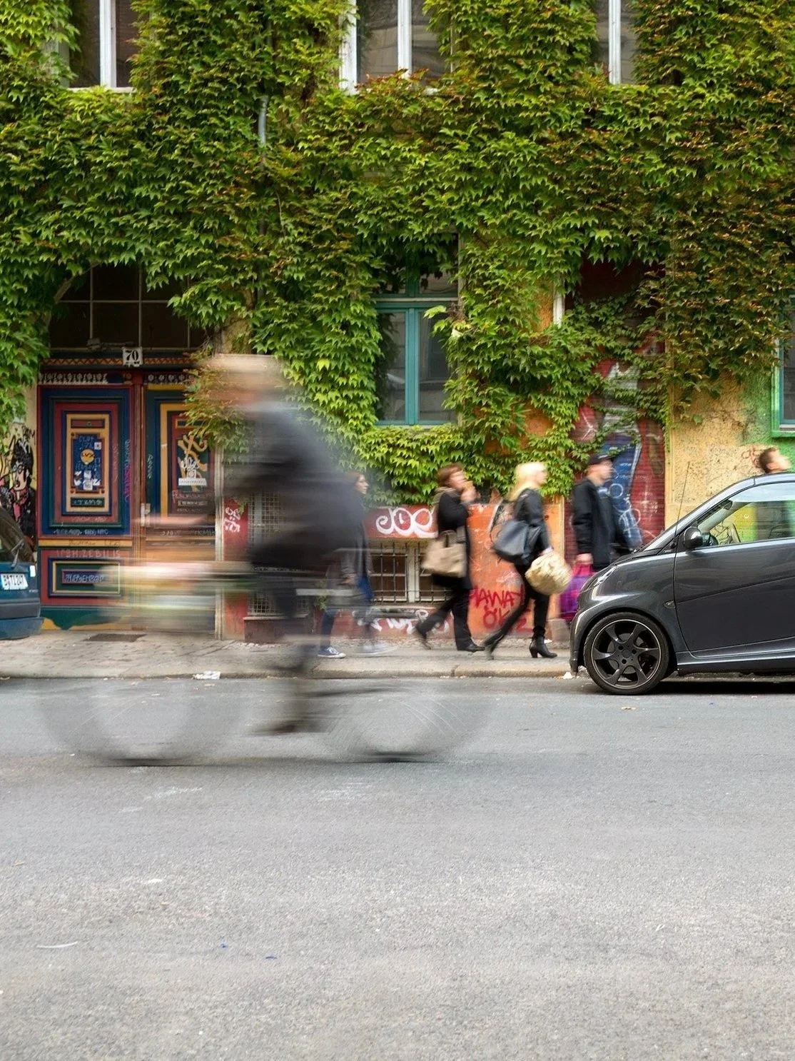 People walking on a city sidewalk with blurred cyclist passing by. Building in the background covered in green ivy, with colorful artwork on the walls.