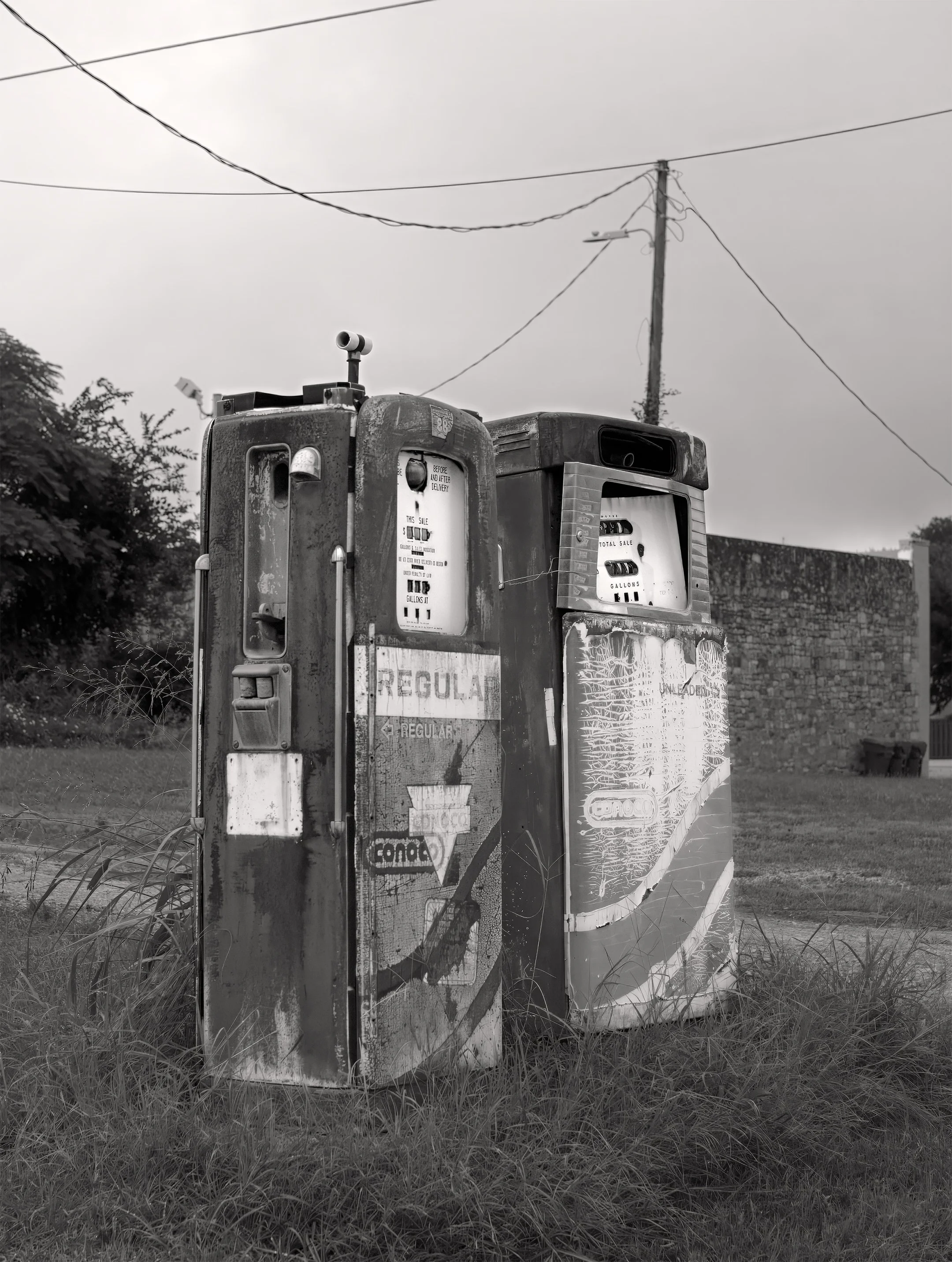Black and white photo of an old, weathered gas pump with a glass globe on top, surrounded by tall grass, with power lines and a utility pole in the background.