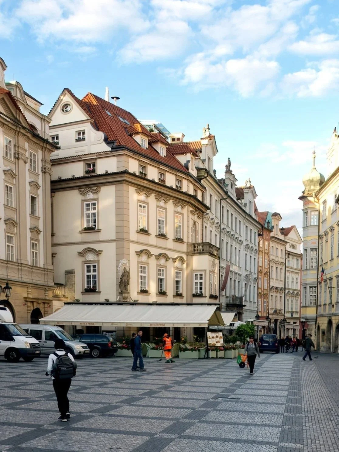 European city square with historic buildings, cobblestone pavement, and people walking, some in casual clothes and others in uniforms.