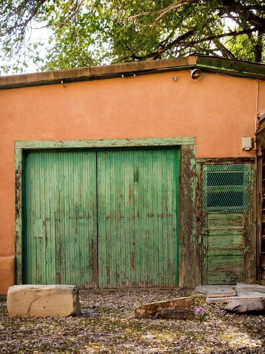 Old wooden green double doors on a building with faded paint, peeling wood, and a smaller window with metal mesh to the right, behind rocks and gravel.