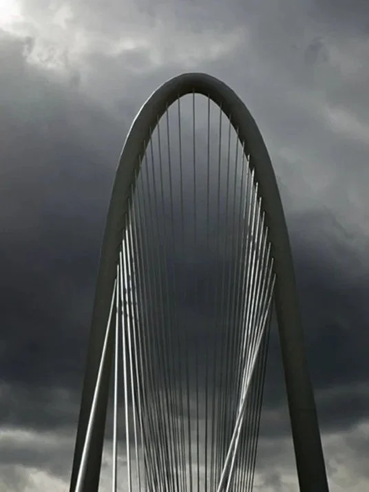 Close-up of a modern, curved suspension bridge with cables against a cloudy, dark sky.