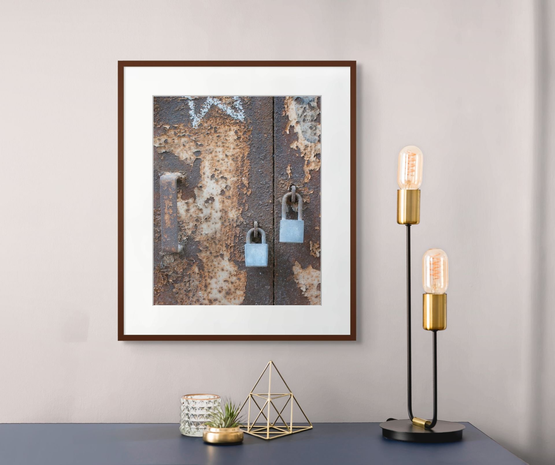 Wall art featuring rusty metal door with padlocks, mounted on light-colored wall next to a modern table lamp with exposed bulb, gold accents, and decorative objects including a glass jar, a gold bowl, and geometric gold decor.
