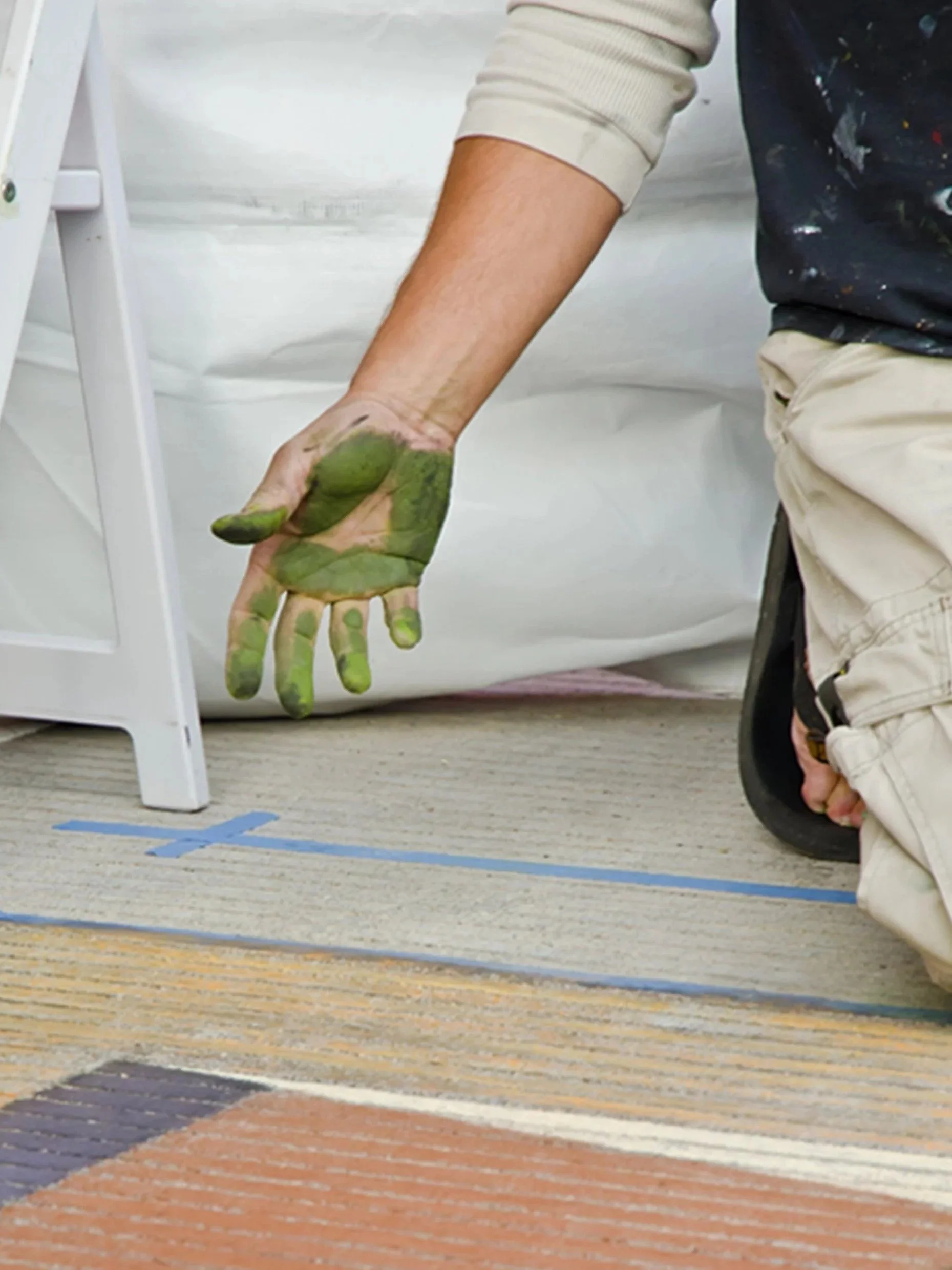 Person kneeling on carpet with green paint on their hand, wearing beige pants and a dark shirt with paint splatters, next to a white table.