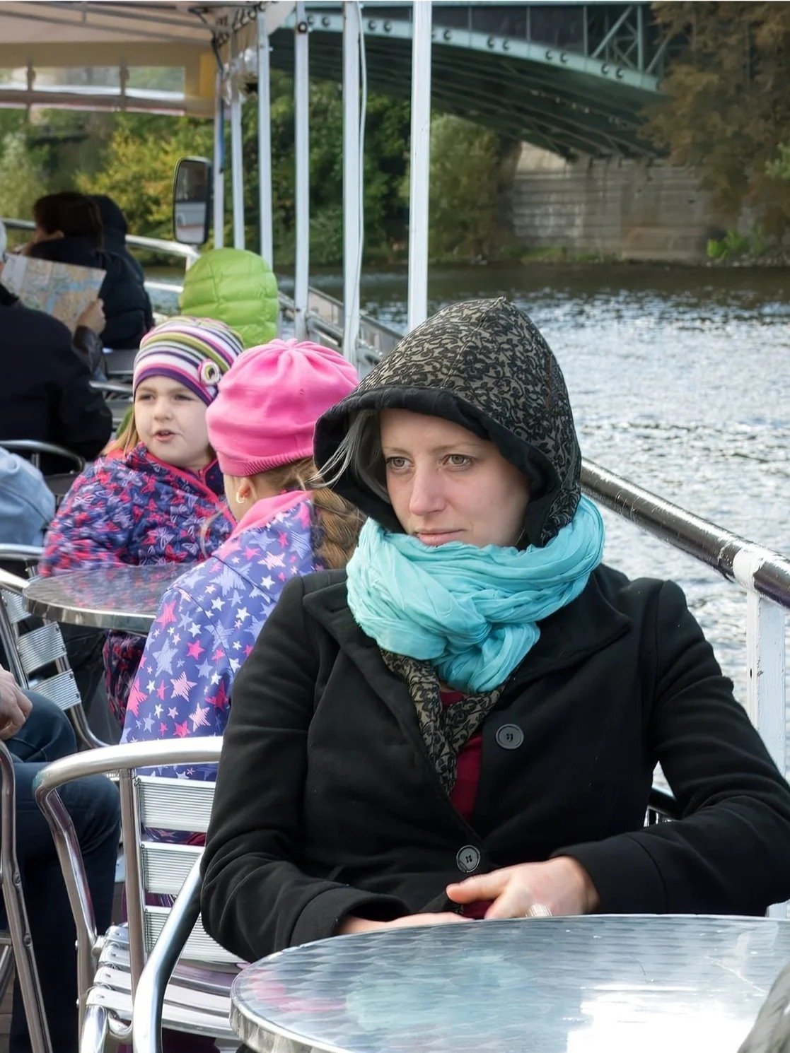A woman wearing a black coat, a black patterned hood, and a turquoise scarf, sitting at a table on a boat, with children in colorful jackets and hats in the background.