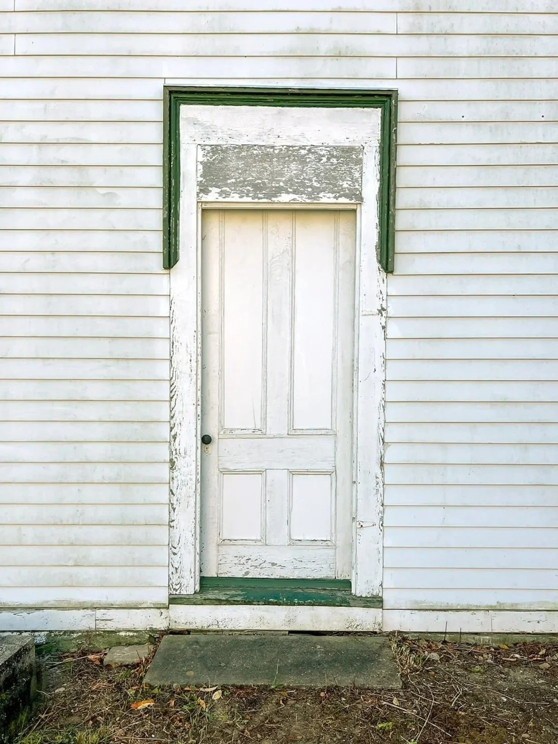 Old weathered white wooden door with peeling paint, set in a white wooden wall with horizontal siding, a small green and gray step in front.
