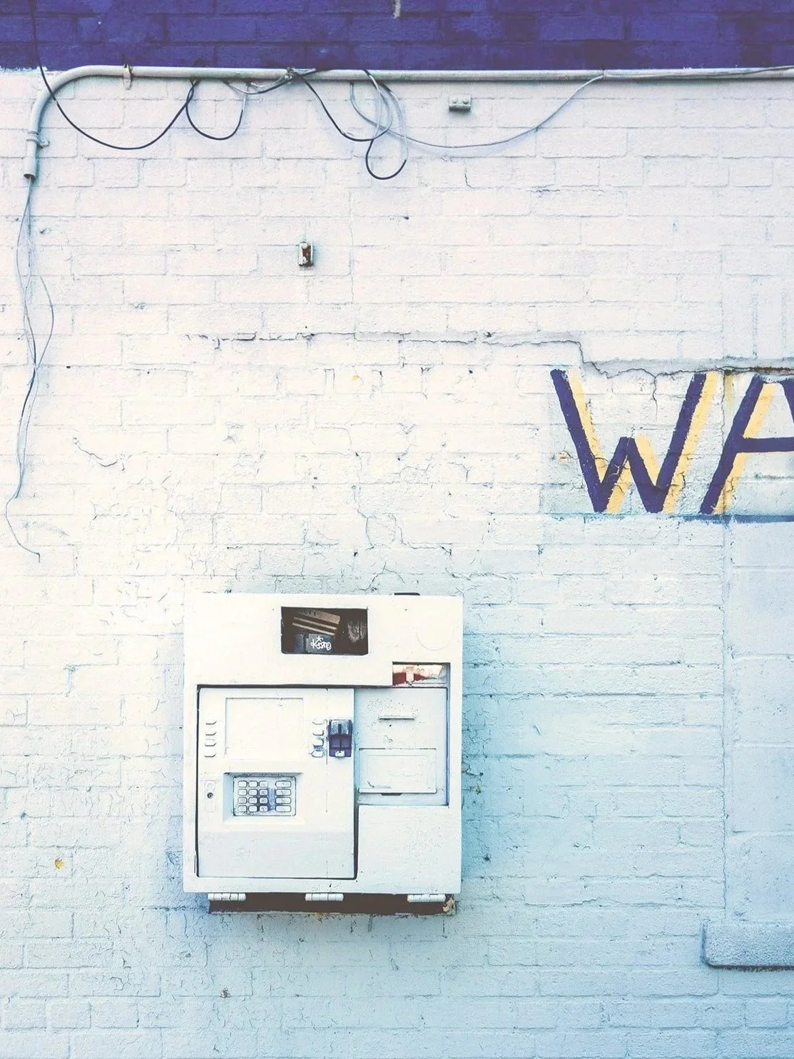 A white vending machine mounted on a white brick wall, with some wires hanging above and partial purple and yellow graffiti on the wall.