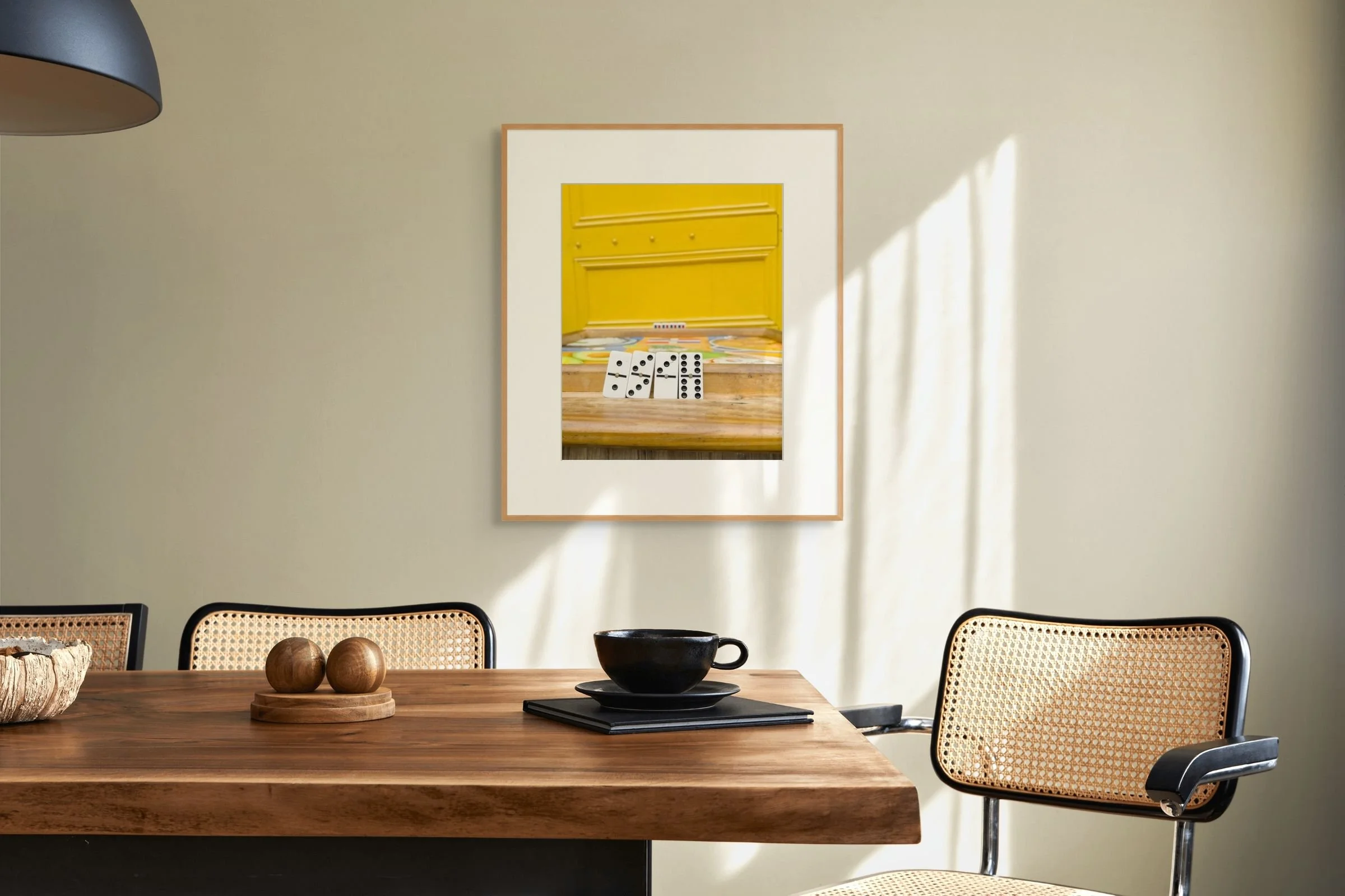 A wooden dining table with three wooden spheres on a round wooden tray, a black cup and saucer, and some books, surrounded by wicker and black chairs, with a framed mirror reflecting the yellow interior of a cabinet and dominoes on a wooden surface in the background.