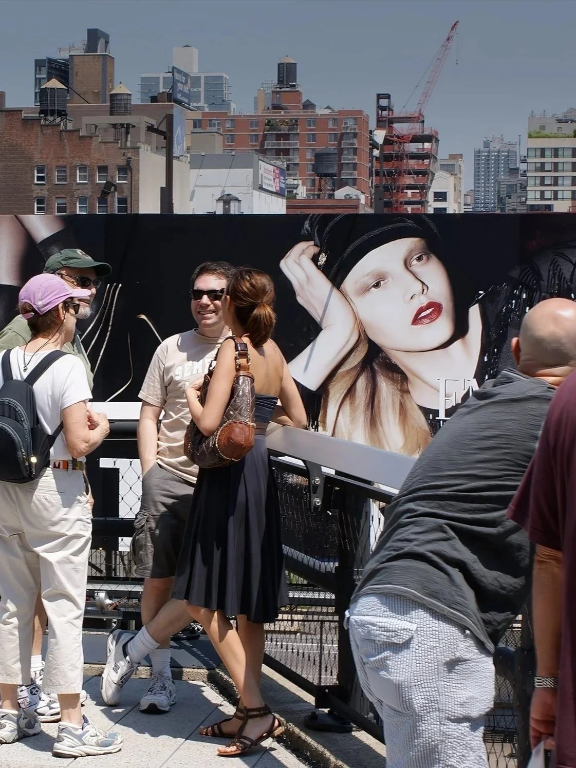 People standing and talking outdoors in front of a large billboard featuring a close-up of a woman with red lipstick and black headscarf, with city buildings and cranes in the background.