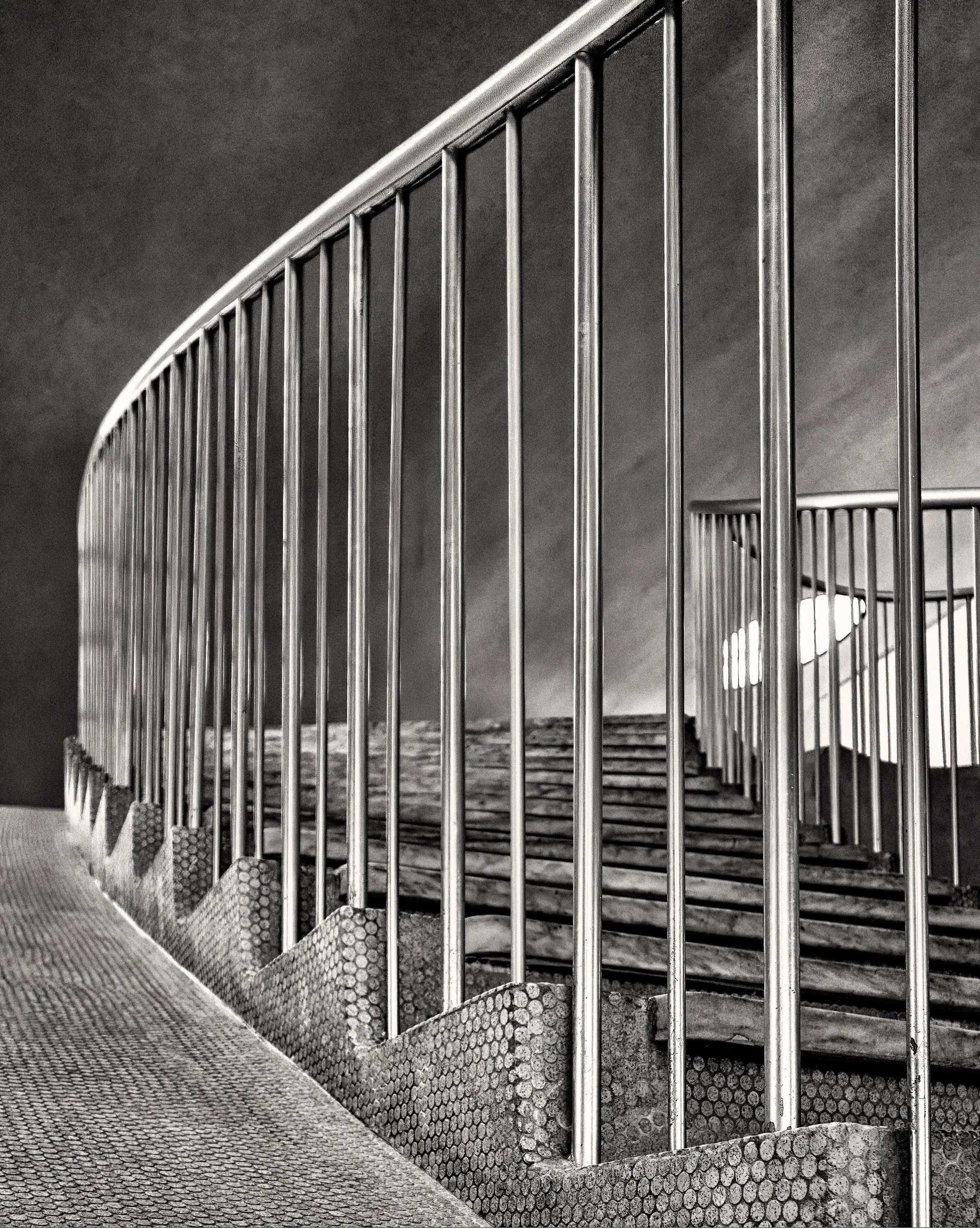 Black and white photo of a metal railing and outdoor staircase with a cloudy sky background.