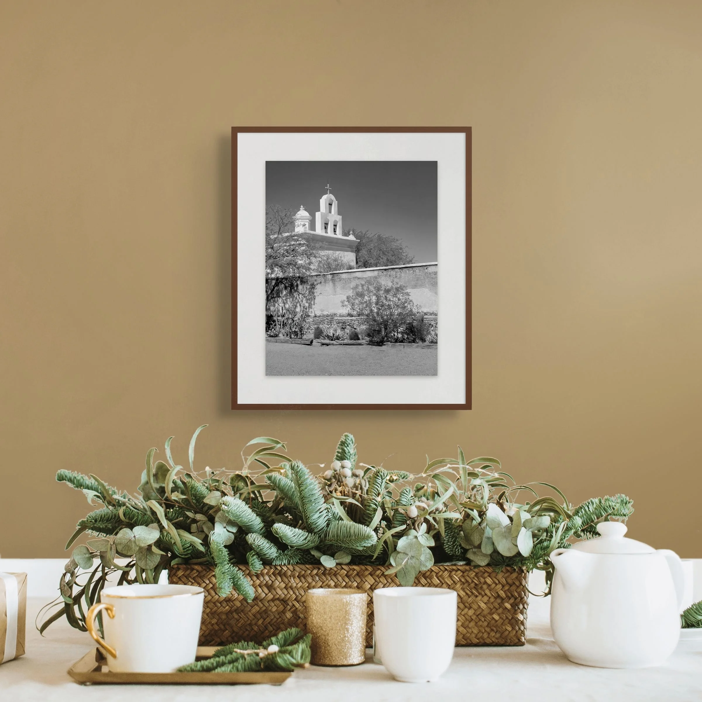 Decorative arrangement of potted plants, white and gold coffee mugs, and a framed black-and-white photograph of a church with a steeple and cross, on a white surface against a beige wall.