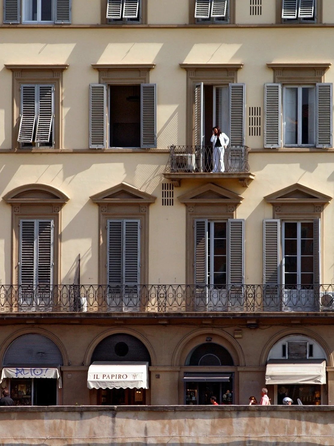 A multi-story building with beige walls, multiple windows with gray shutters. A woman in white stands on a small balcony on the upper floor, looking outside.