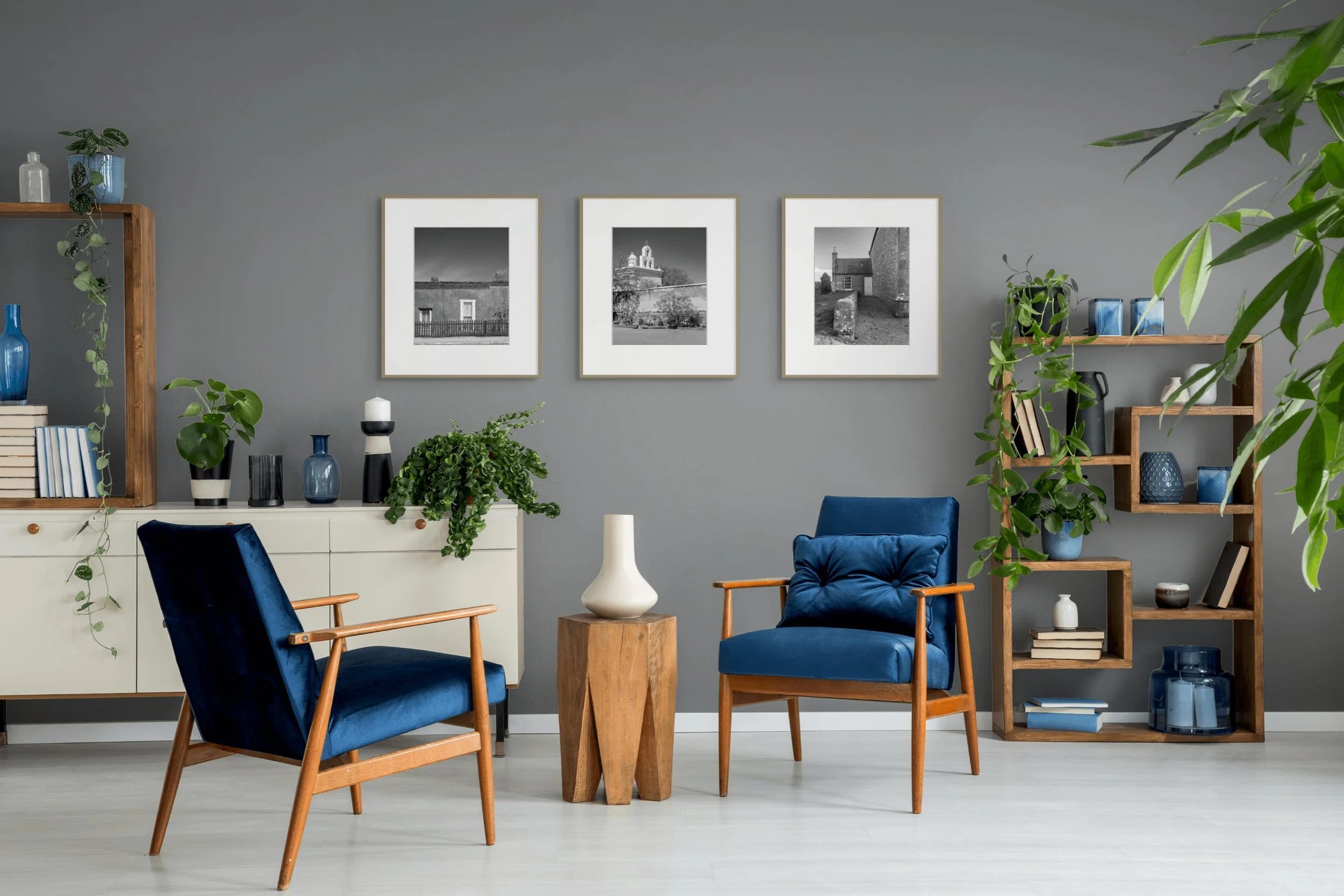 Living room with gray wall, three black-and-white framed photos, wooden shelves with plants and decorative items, blue velvet chairs, and a white sideboard.