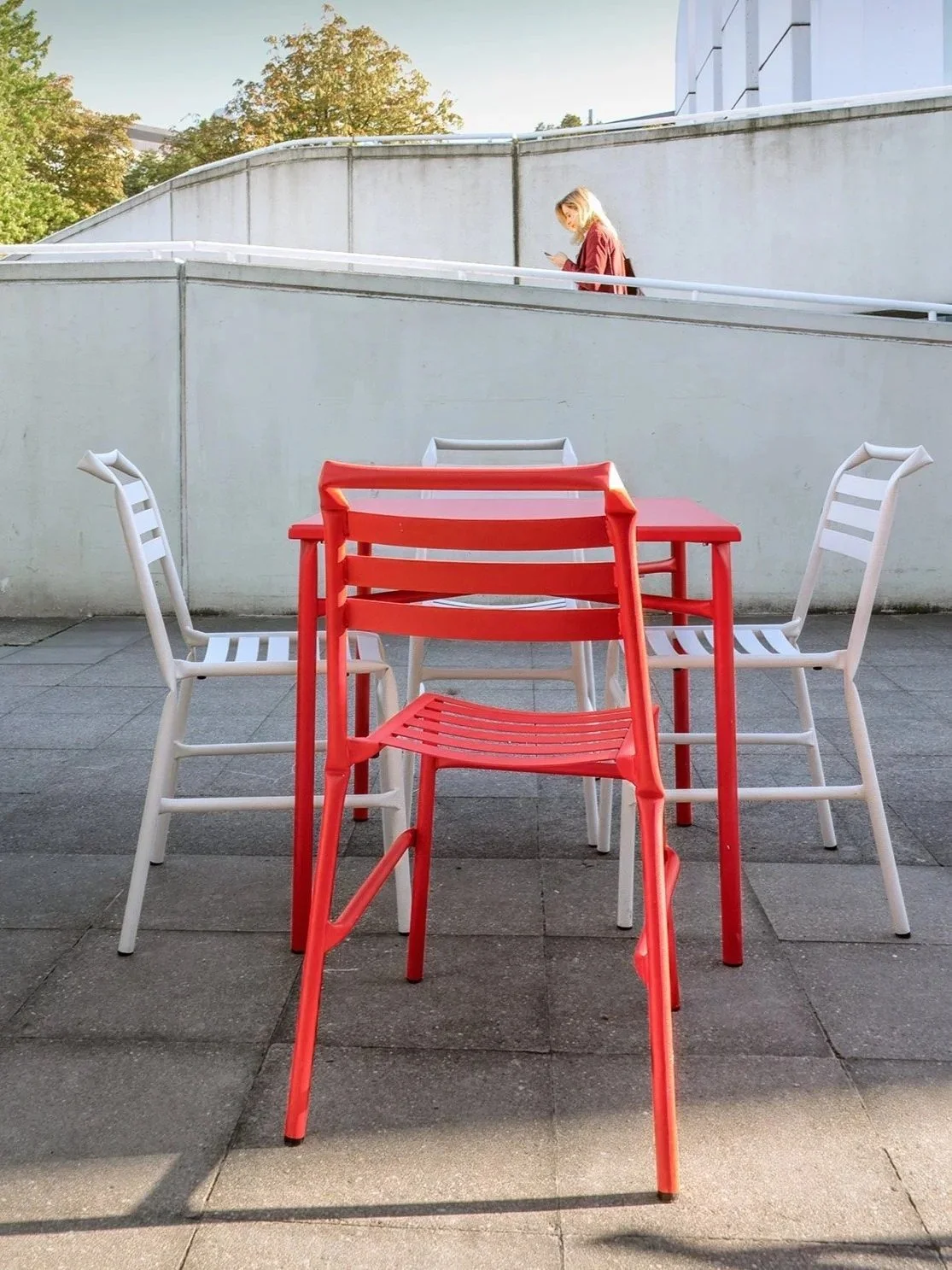 A red chair and a red table surrounded by four white chairs on a paved outdoor patio. 