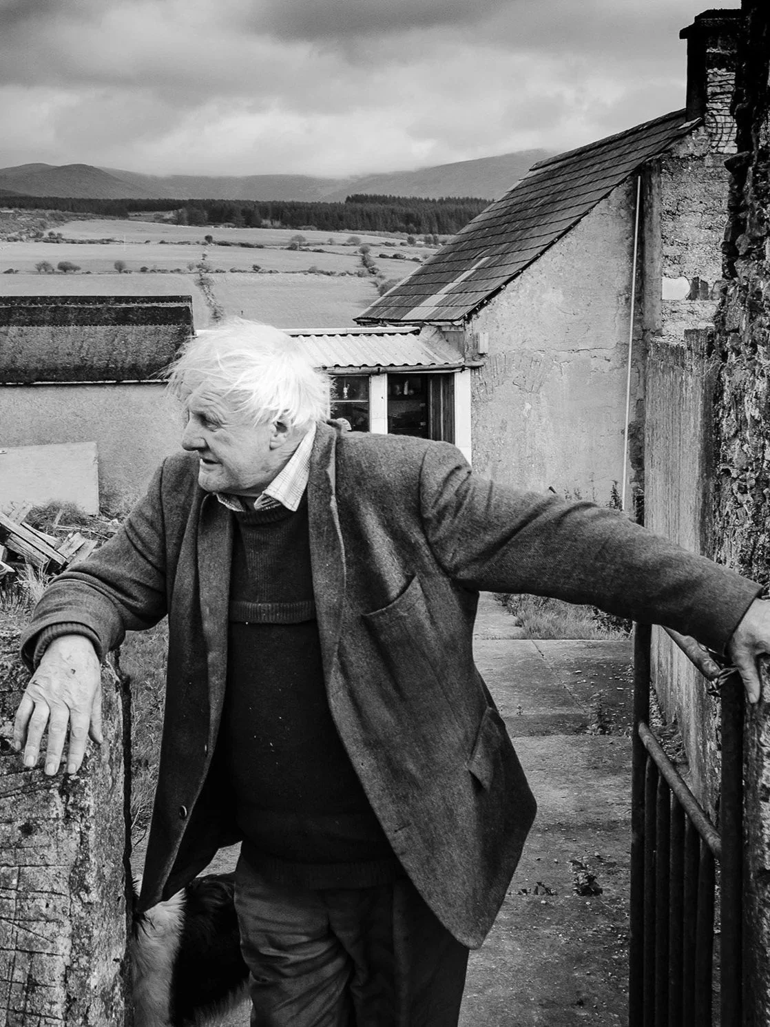 An elderly man with white hair wearing a blazer and sweater, leaning on a stone wall.