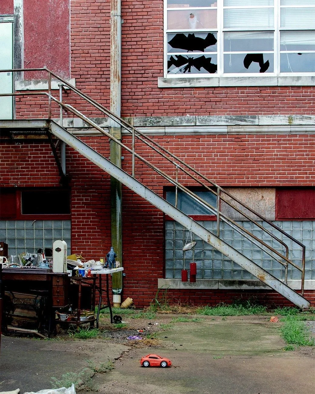 View of a brick building with a metal fire escape staircase, broken window, cluttered table, and a small red toy car on the ground.