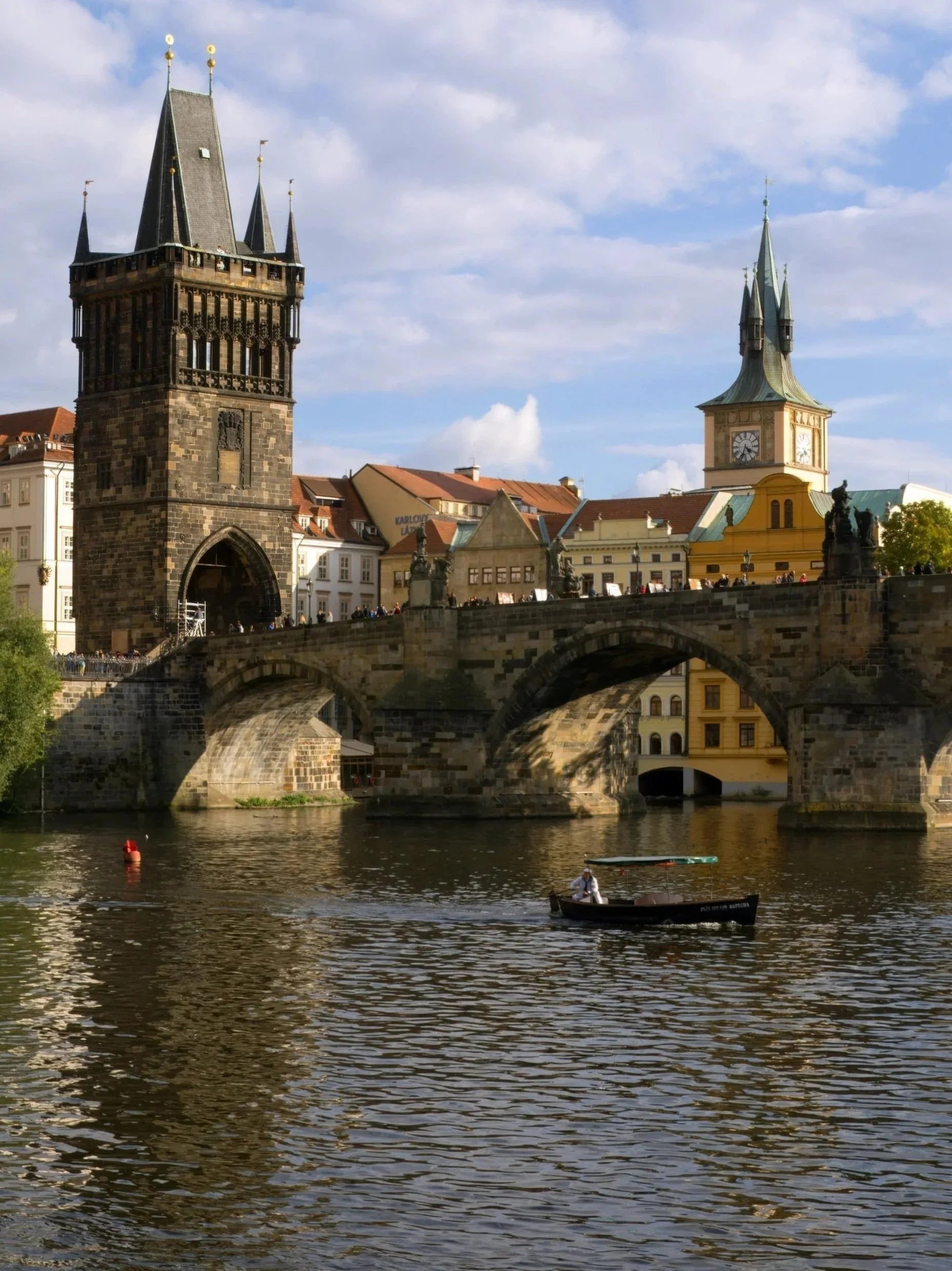View of a historic stone bridge over a river with a boat in the foreground. In the background, there are two tall, ornate clock towers and colorful buildings under a partly cloudy sky.