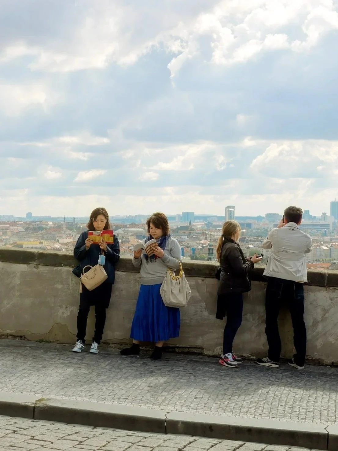 Four people standing on a balcony or observation deck, looking out over a city skyline with clouds in the sky.