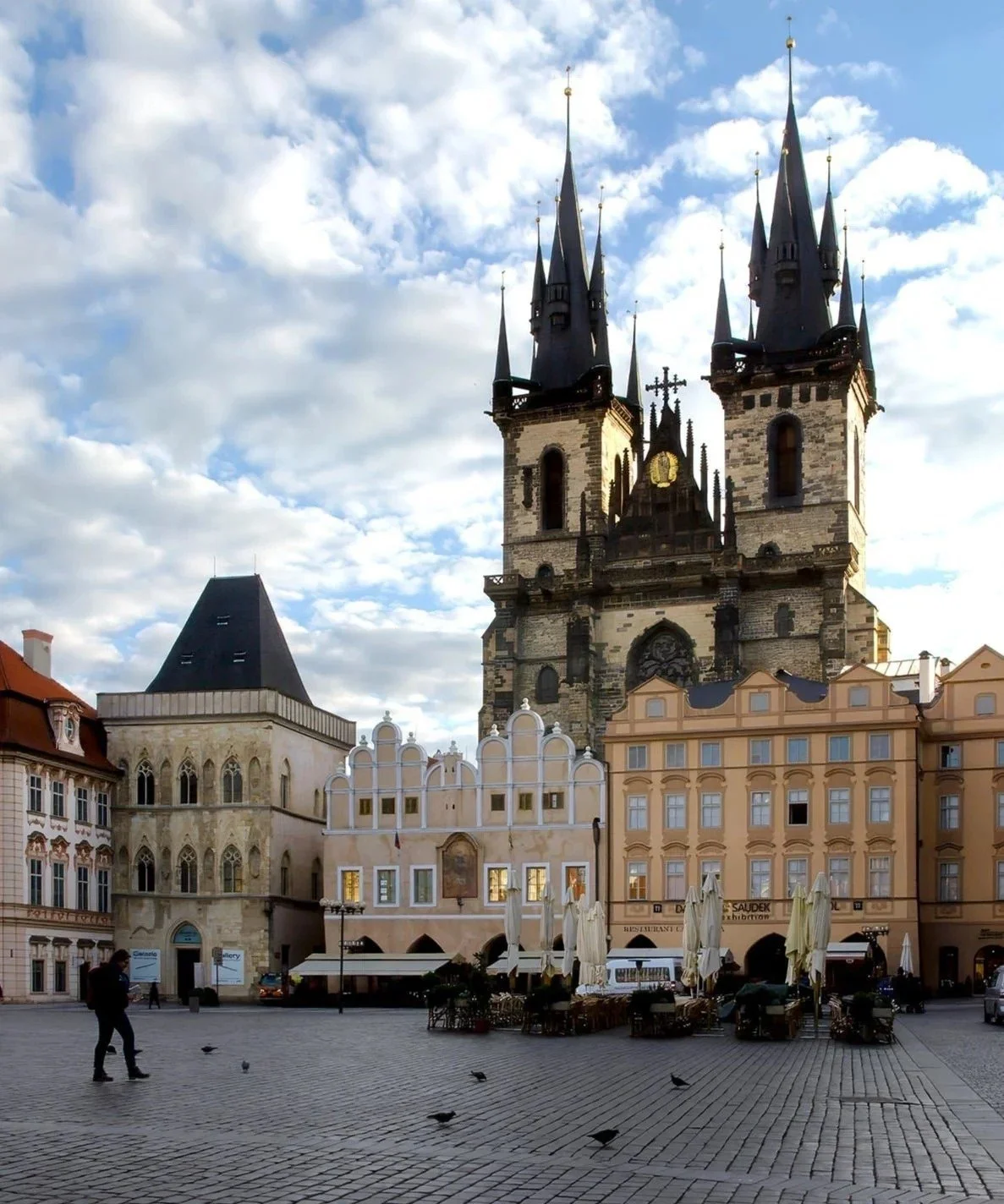 View of a historic European square with colorful buildings, outdoor seating, and a large Gothic church with twin towers in the background, under a partly cloudy sky.