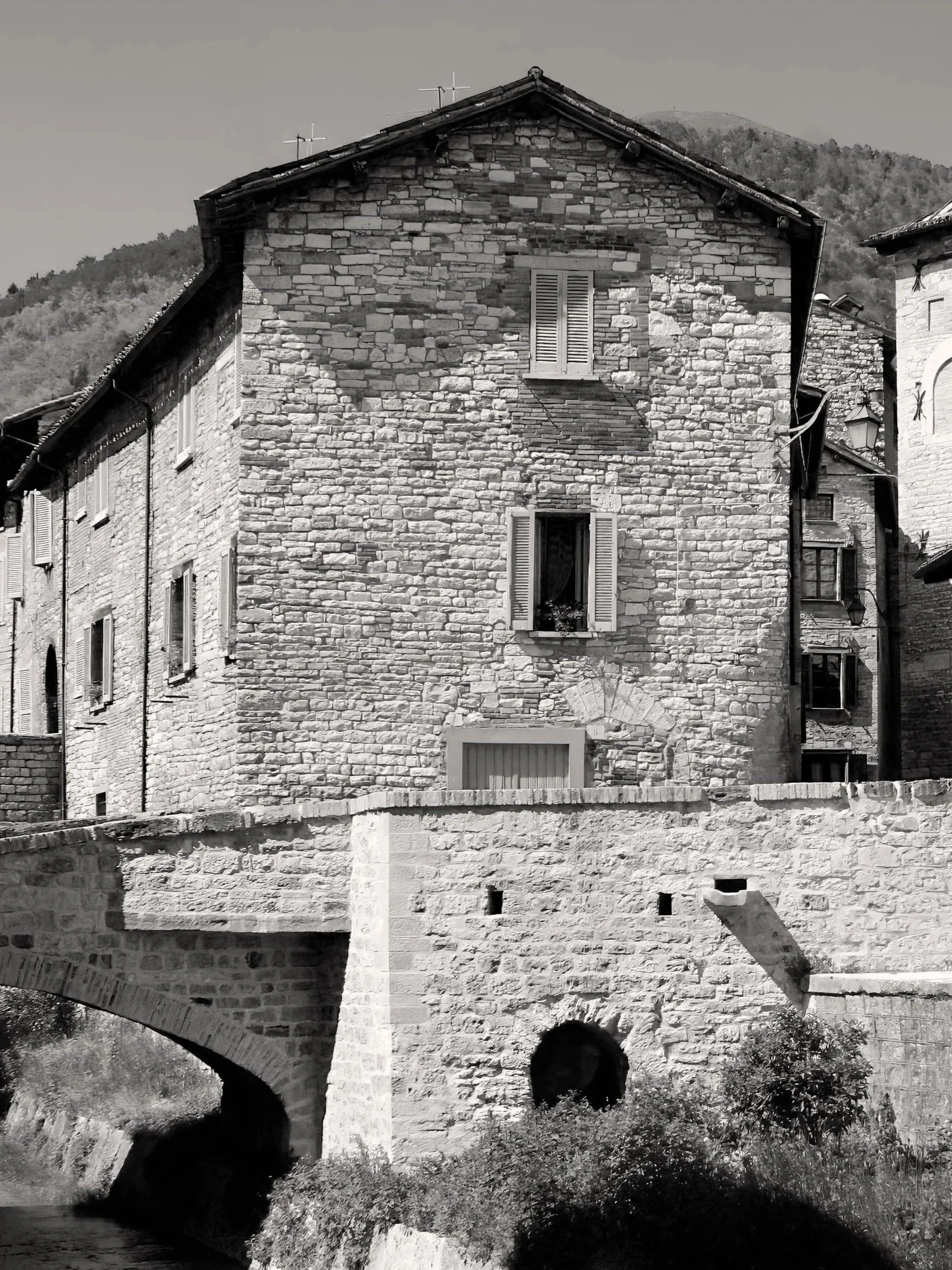 A black and white photograph of a stone building with shutters on the windows, situated next to a small bridge over a creek.