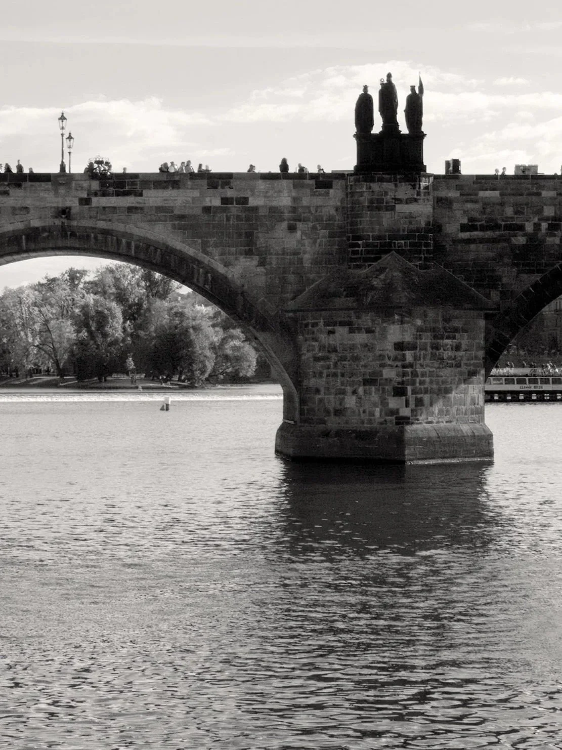 Black and white photo of a stone bridge over a river, featuring a statue on top of the bridge, with a few people walking along the bridge, trees on the riverbank, and a boat on the water.