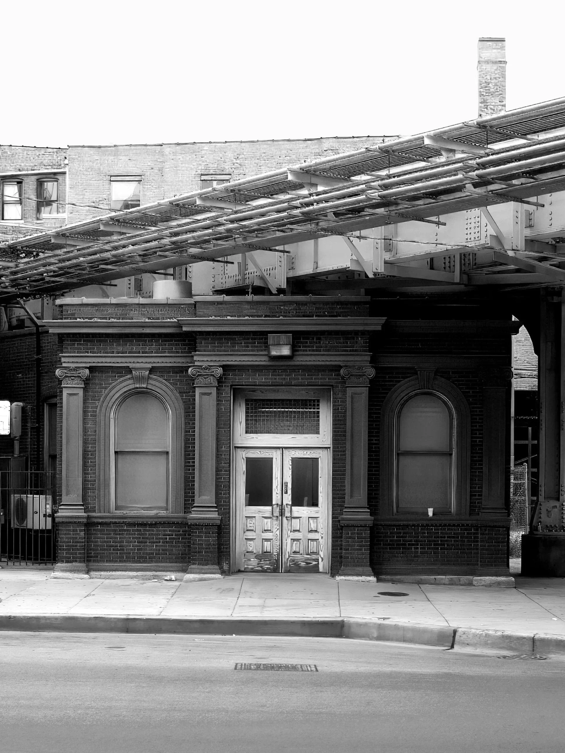 Black and white photo of an old brick building with ornate window frames and a pair of updating doors. A metallic overhead structure is above, with a city sidewalk and street in front.
