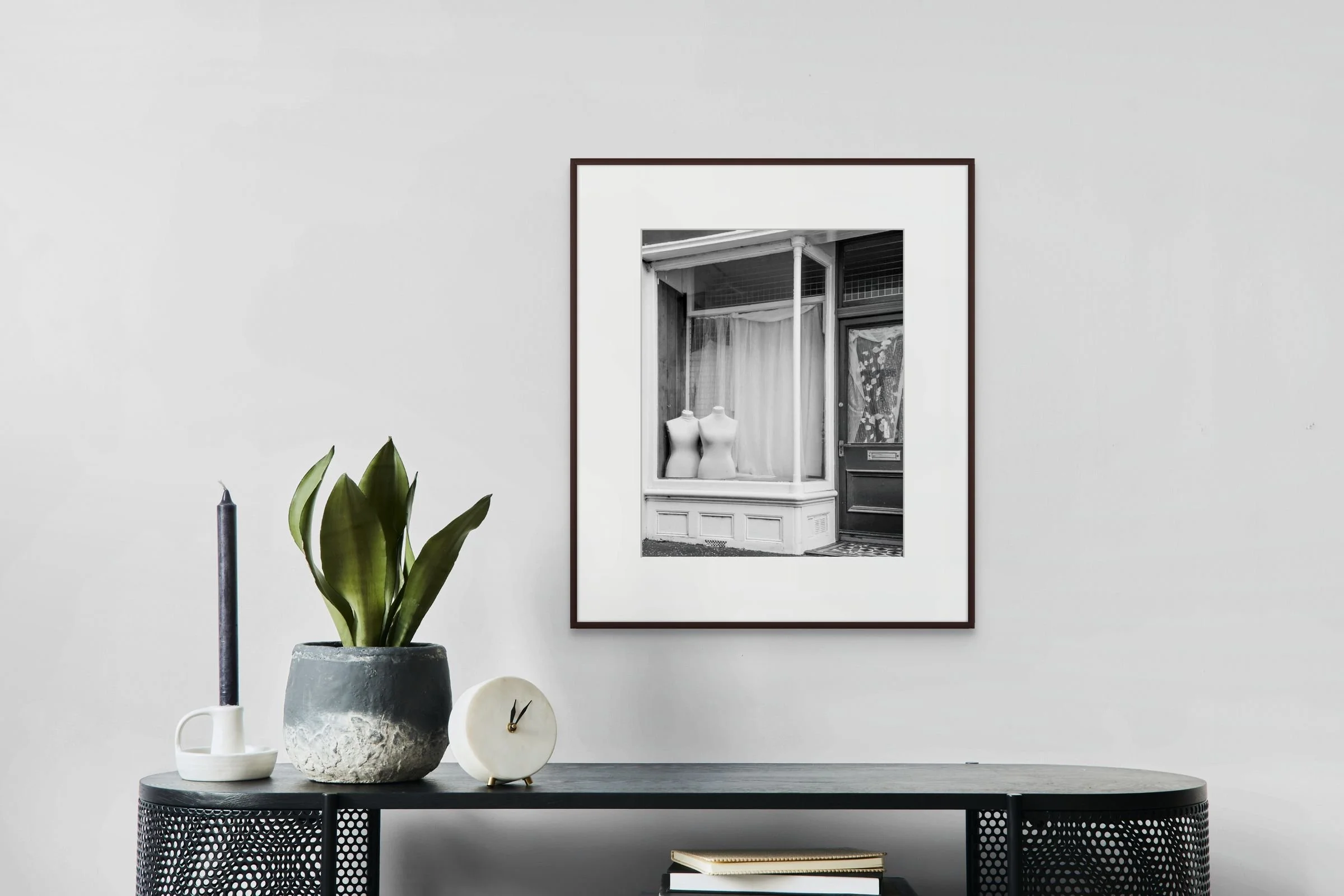 Decorative setup on a black table with a potted plant, a small clock, a mug, and books, with a framed black-and-white photograph hanging on a plain white wall behind.