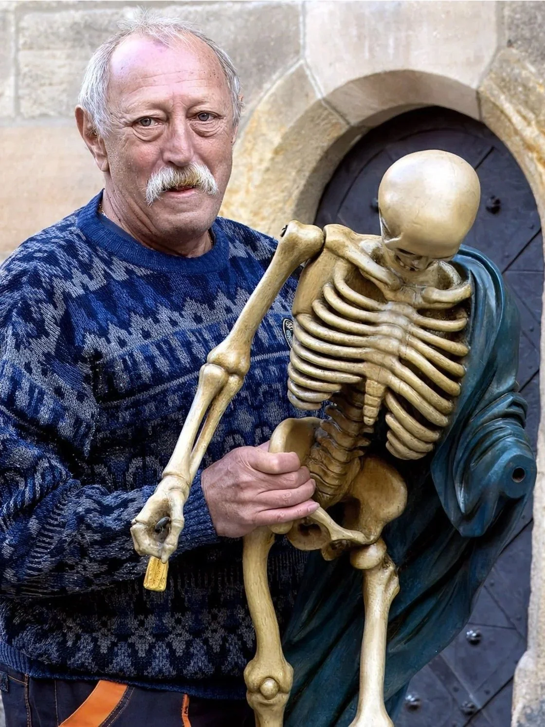 An elderly man with a mustache and wearing a blue patterned sweater holding a human skeleton model in a museum.