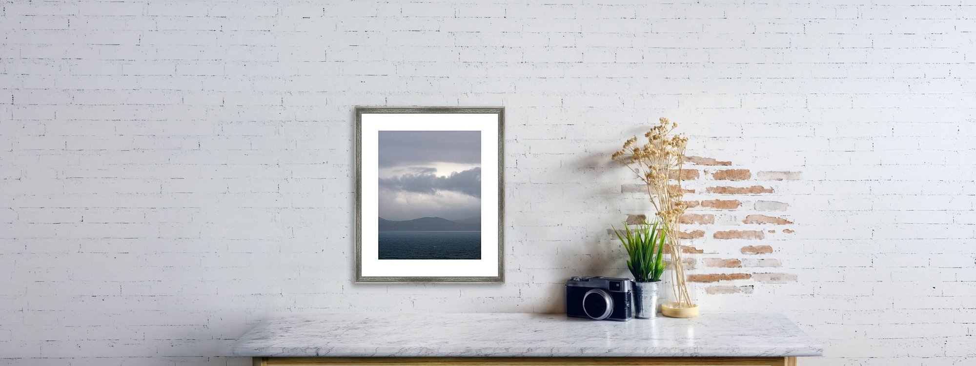 Decorative table against white brick wall with a framed landscape photo, a camera, a potted plant, and dried flowers.