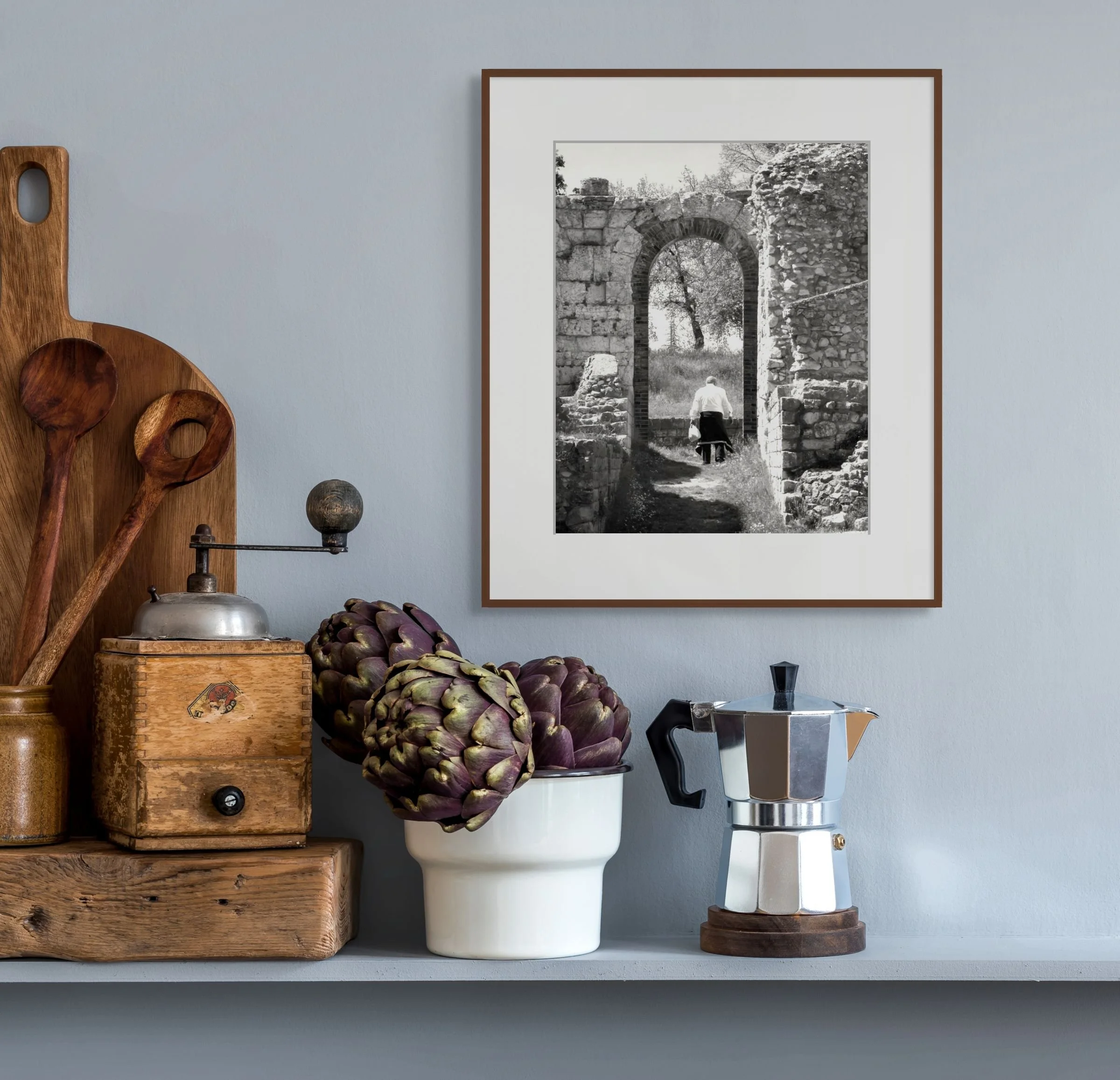 A wall with three framed black-and-white photos of ancient stone ruins, a wooden shelf holding wooden kitchen utensils and a vintage wooden box, and artichokes on a surface, with a moka pot coffee maker.