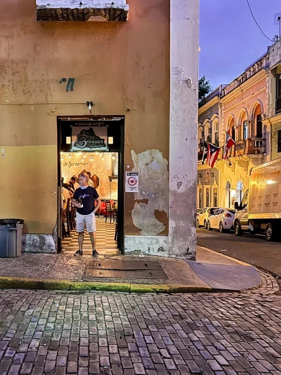 A man standing outside the entrance of a restaurant on a cobblestone street during early evening, with several parked cars and European-style buildings with flags in the background.