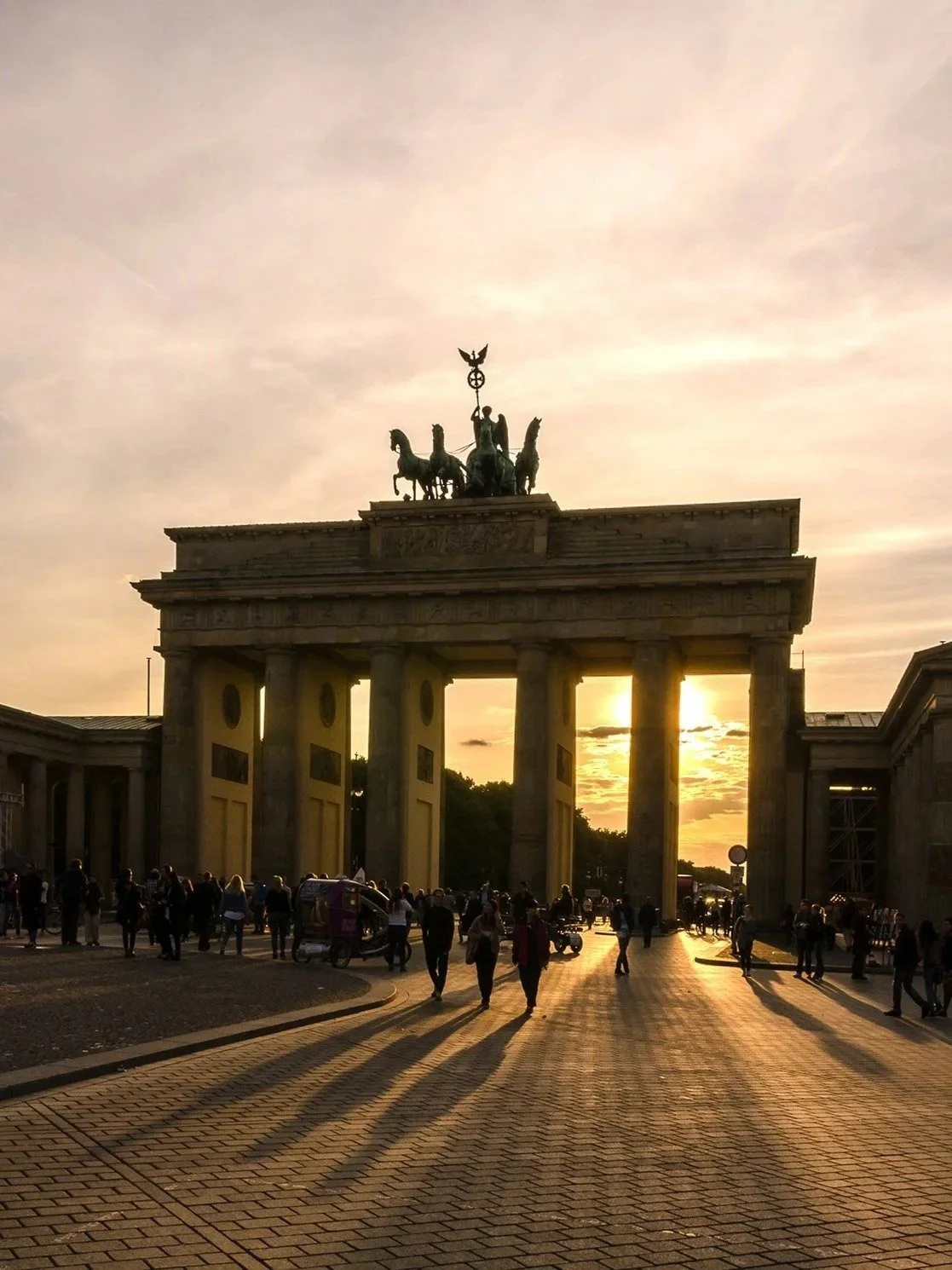 Sunset view of the Brandenburg Gate in Berlin, Germany, with people walking and shadows cast on the street.