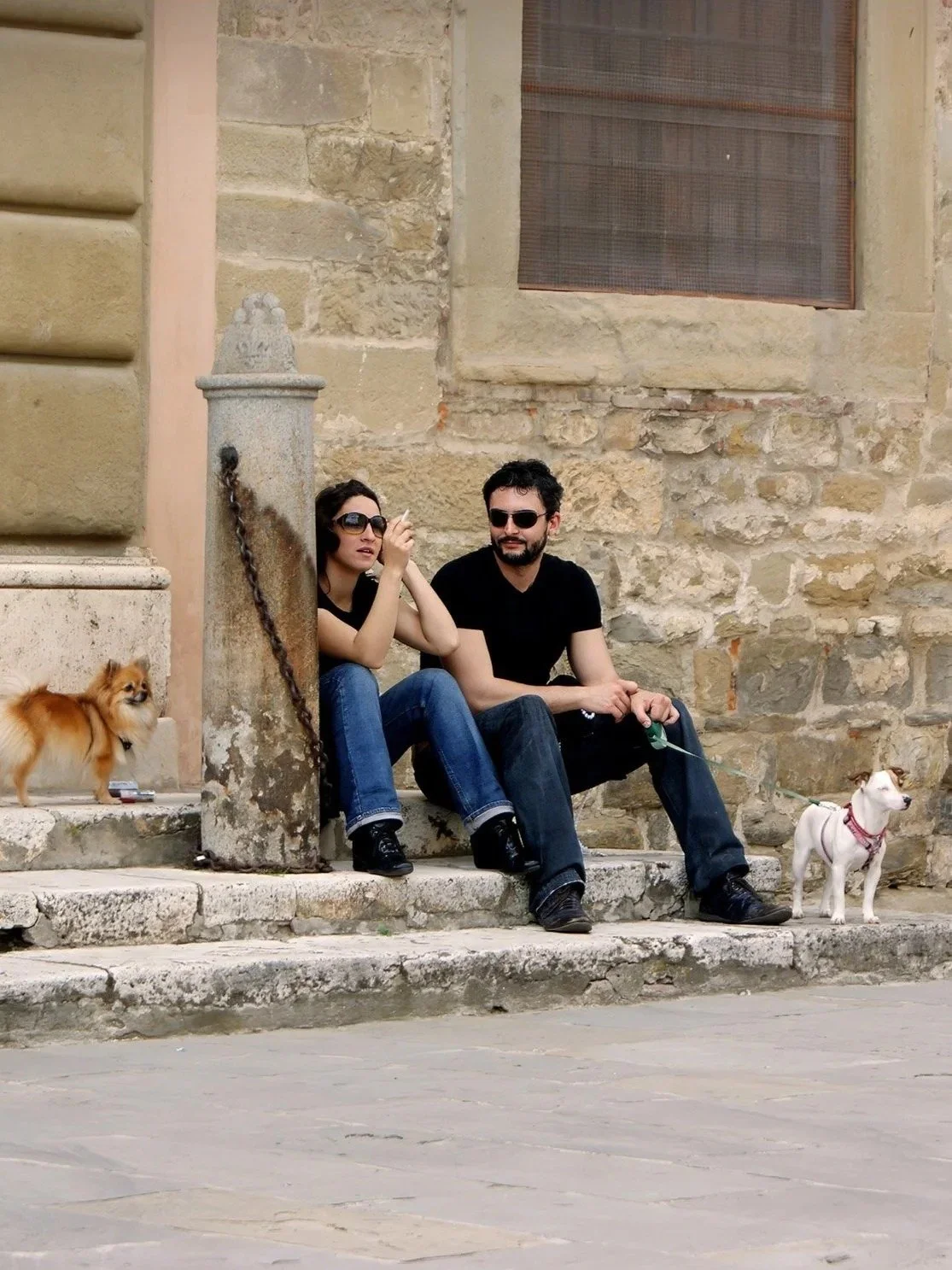 A man and a woman sitting on stone steps outside a building, each holding a dog on a leash.