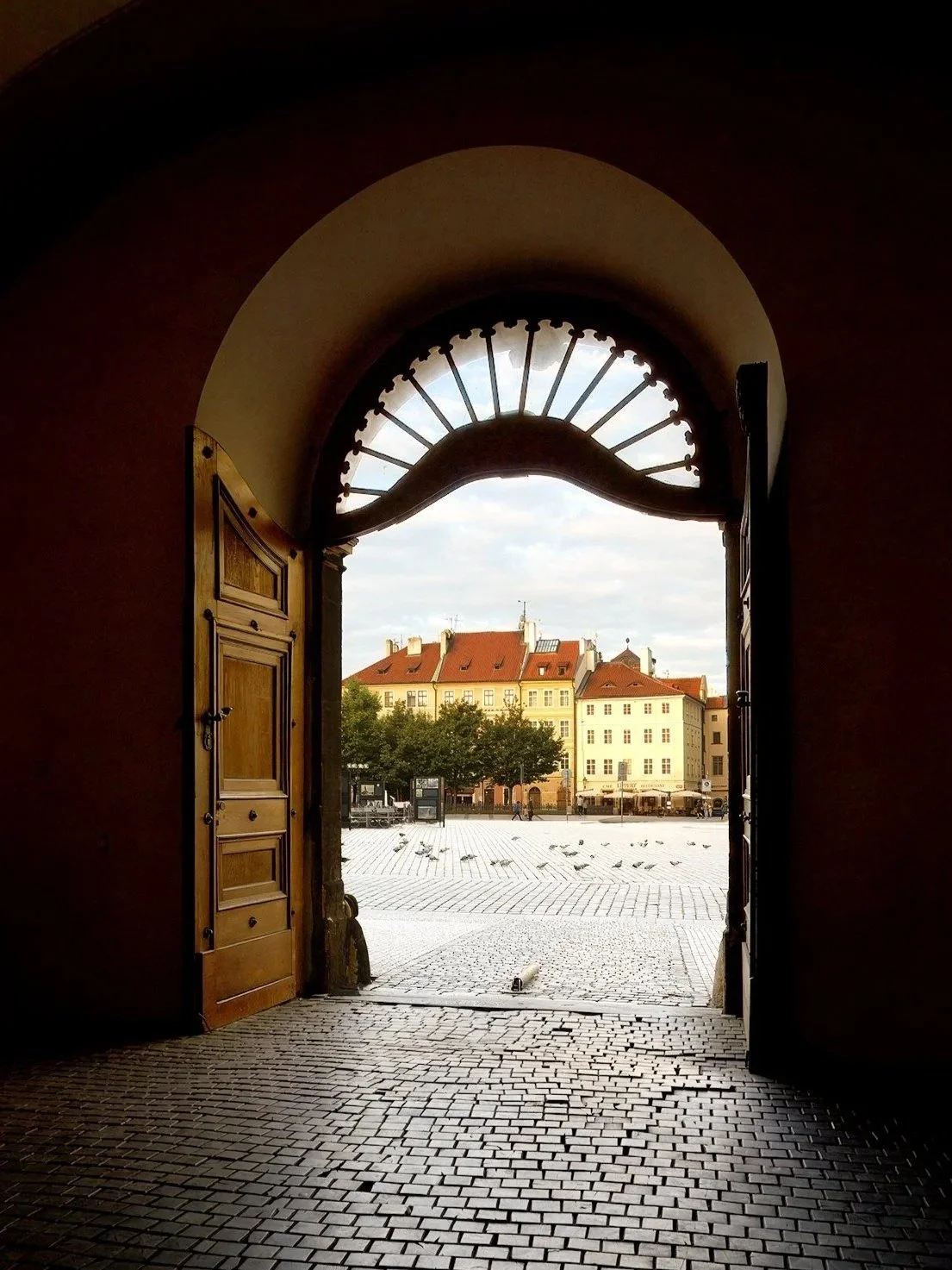 View of a city square seen through an open arched doorway with wooden doors in a historic building.